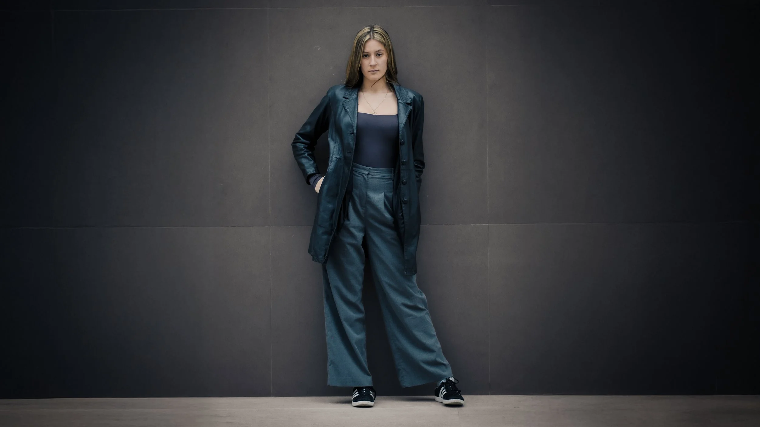 Ragazza giovane con cappotto lungo e pantaloni larghi si appoggia a una parete scura in un locale moderno.