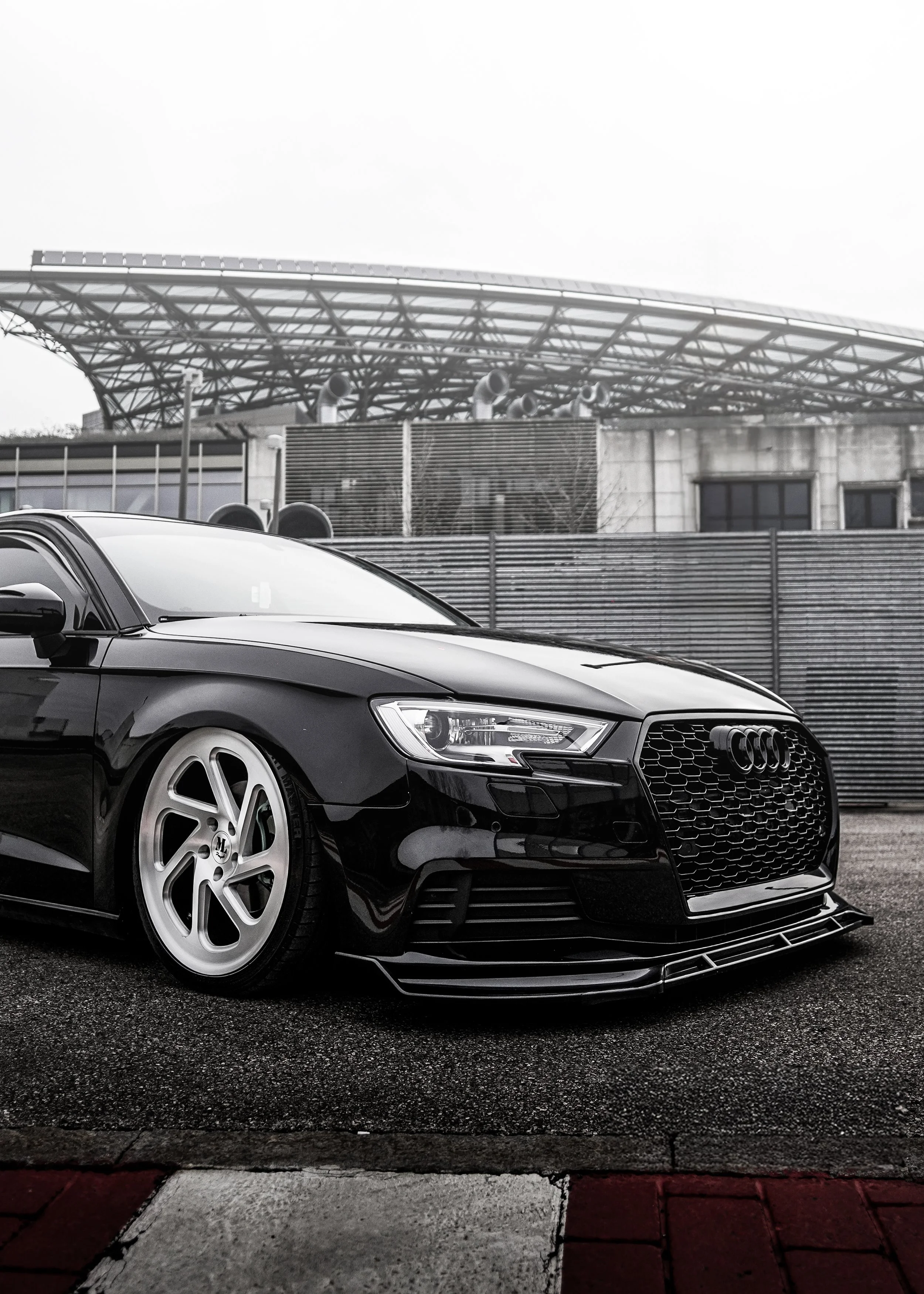 Auto nera Audi con cerchioni bianchi davanti a una recinzione metallica e una struttura moderna sullo sfondo.