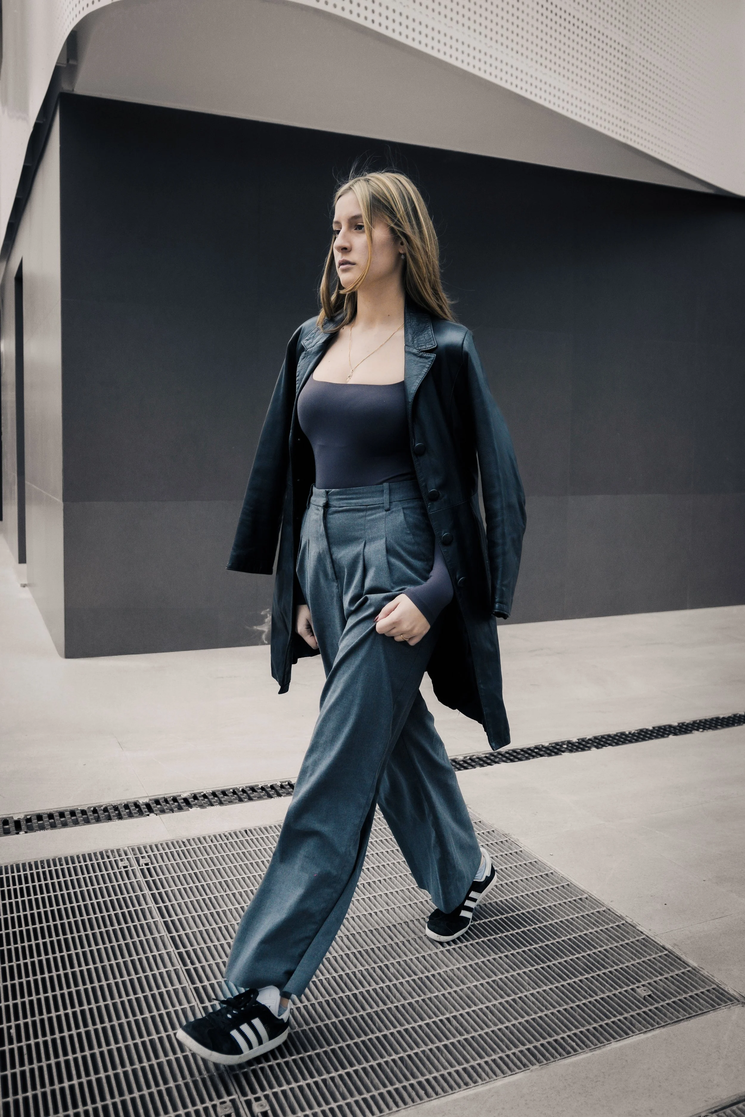 Una donna cammina in un'area moderna, indossa un abbigliamento casual con giacca di pelle, top scollato, pantaloni larghi e scarpe da ginnastica, con edifici moderni sullo sfondo.