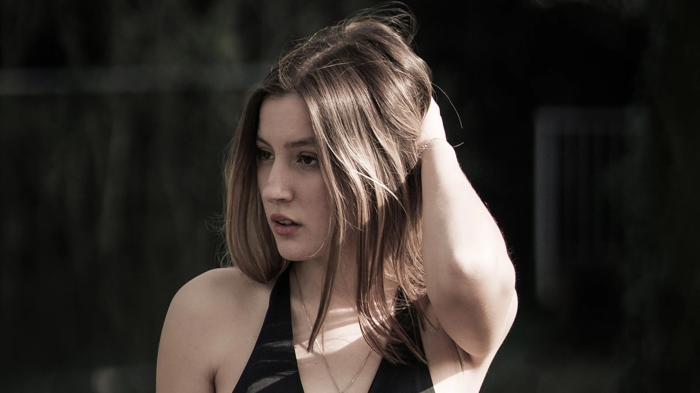 Giovane donna con capelli castani, vestita con top nero, che si tiene i capelli con la mano destra, ambientata in un contesto esterno con sfondo sfocato.