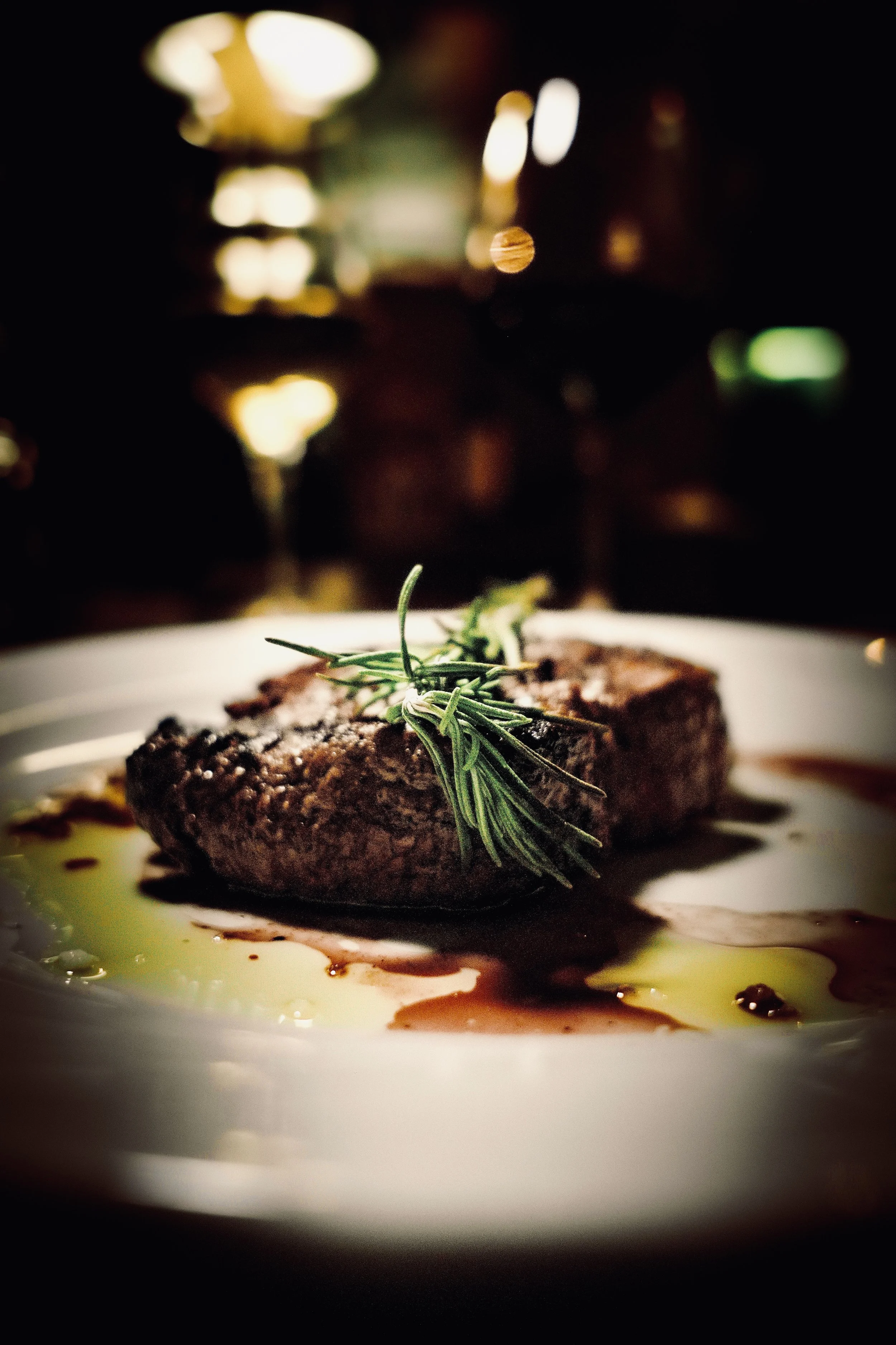 Bistecca di carne con un rametto di rosmarino, servita con oli e salse su un piatto bianco.