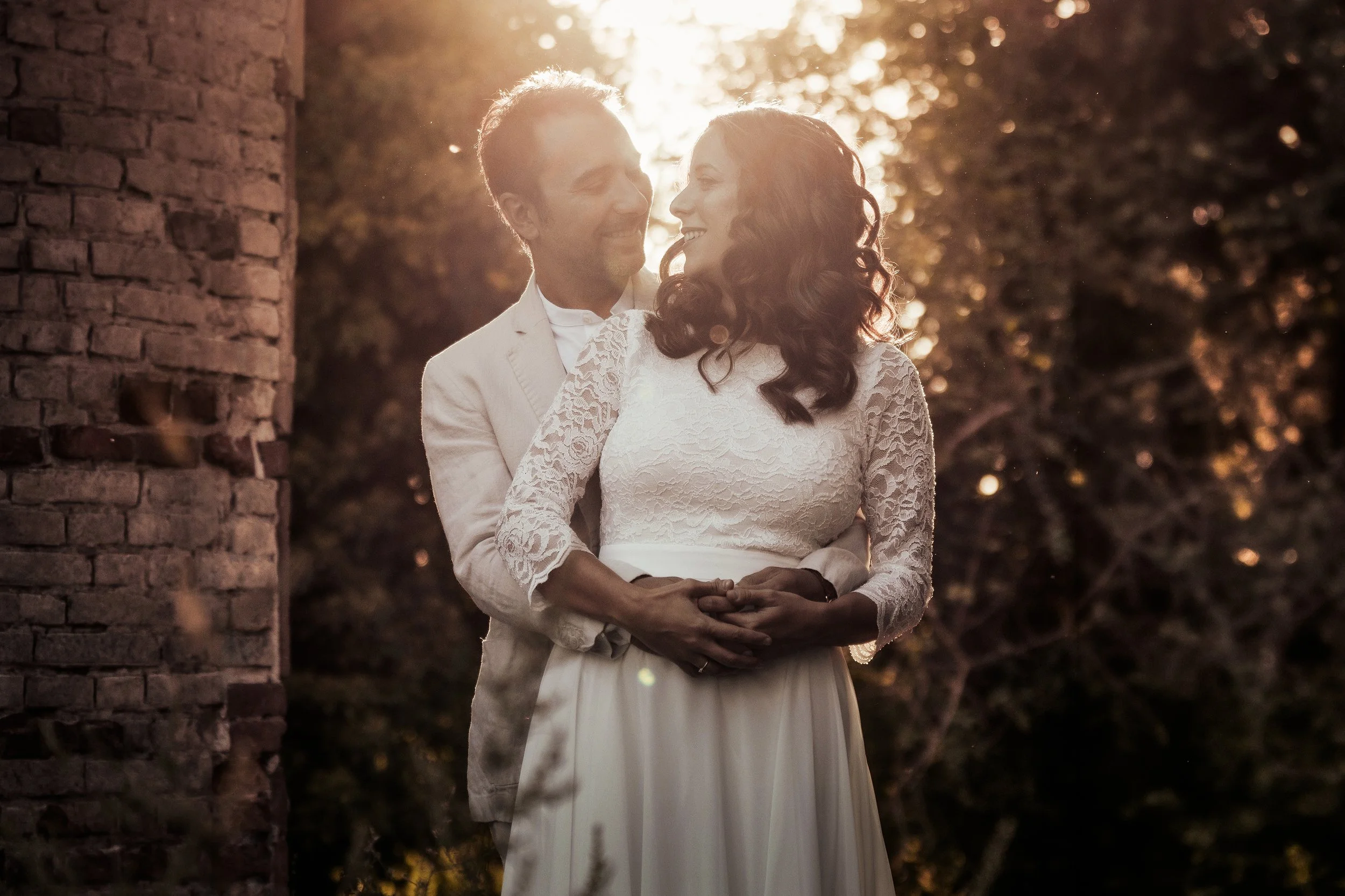 Una coppia di sposi in abiti eleganti si abbracciano e sorridono, in un ambiente naturale al tramonto.