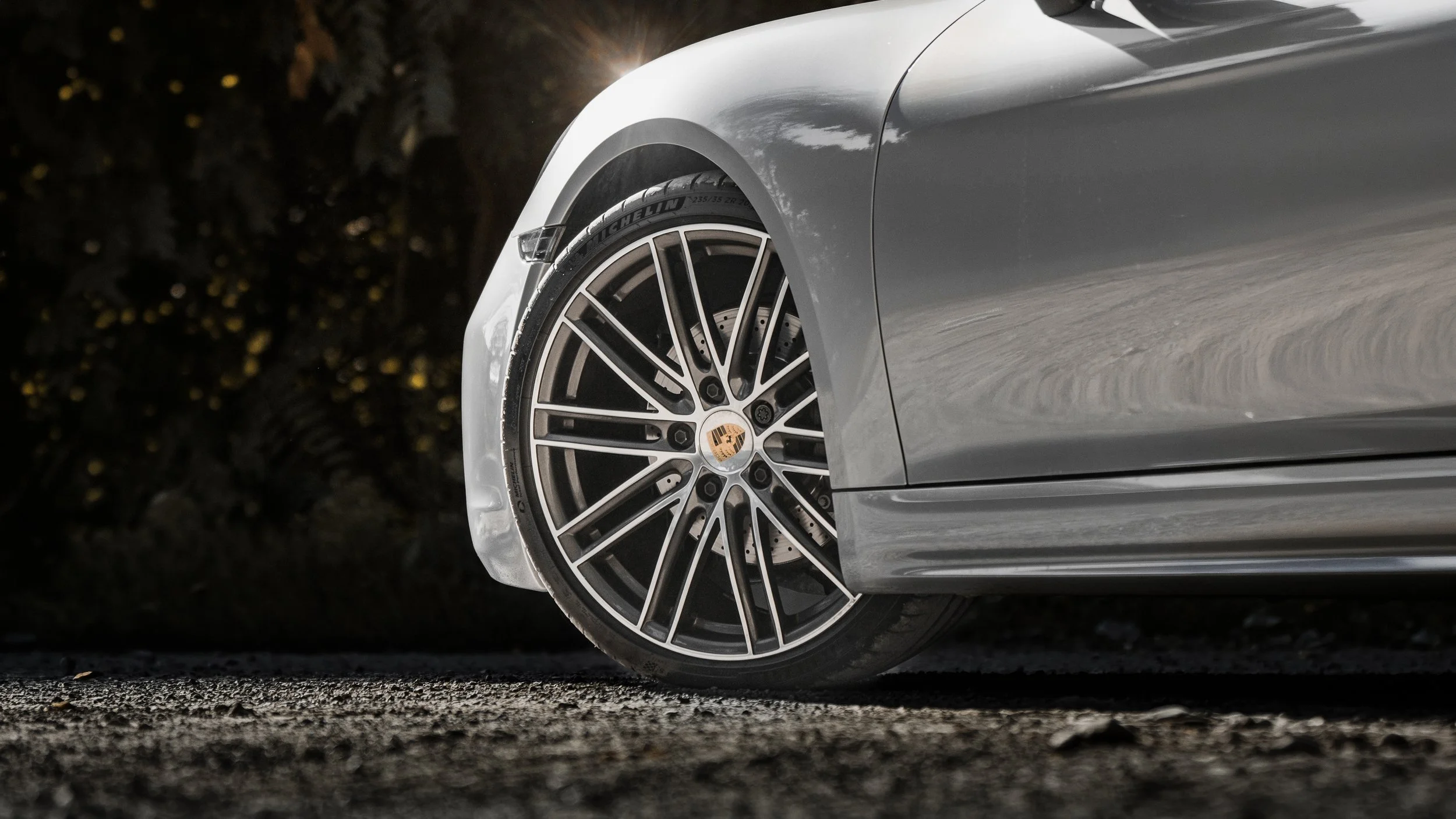 Particolare del lato anteriore di una macchina sportiva grigia con cerchioni sottili e logo Porsche al centro, su strada asfaltata con sfondo di foglie e vegetazione.