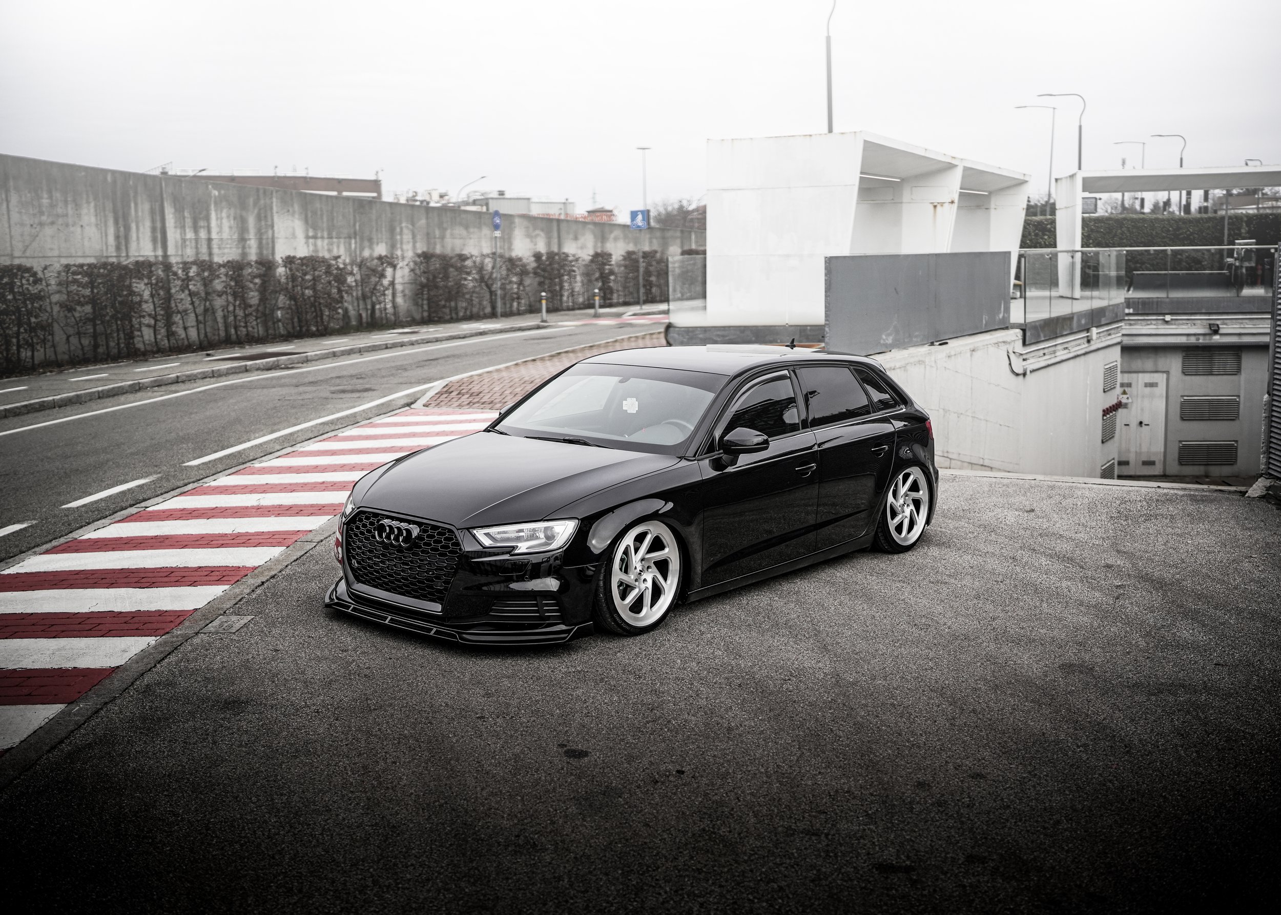 Auto nero di lusso Audi parcheggiata su strada in un'area urbana con sfondo di edifici e una barriera di cemento, cielo grigio e condizioni atmosferiche nuvolose.