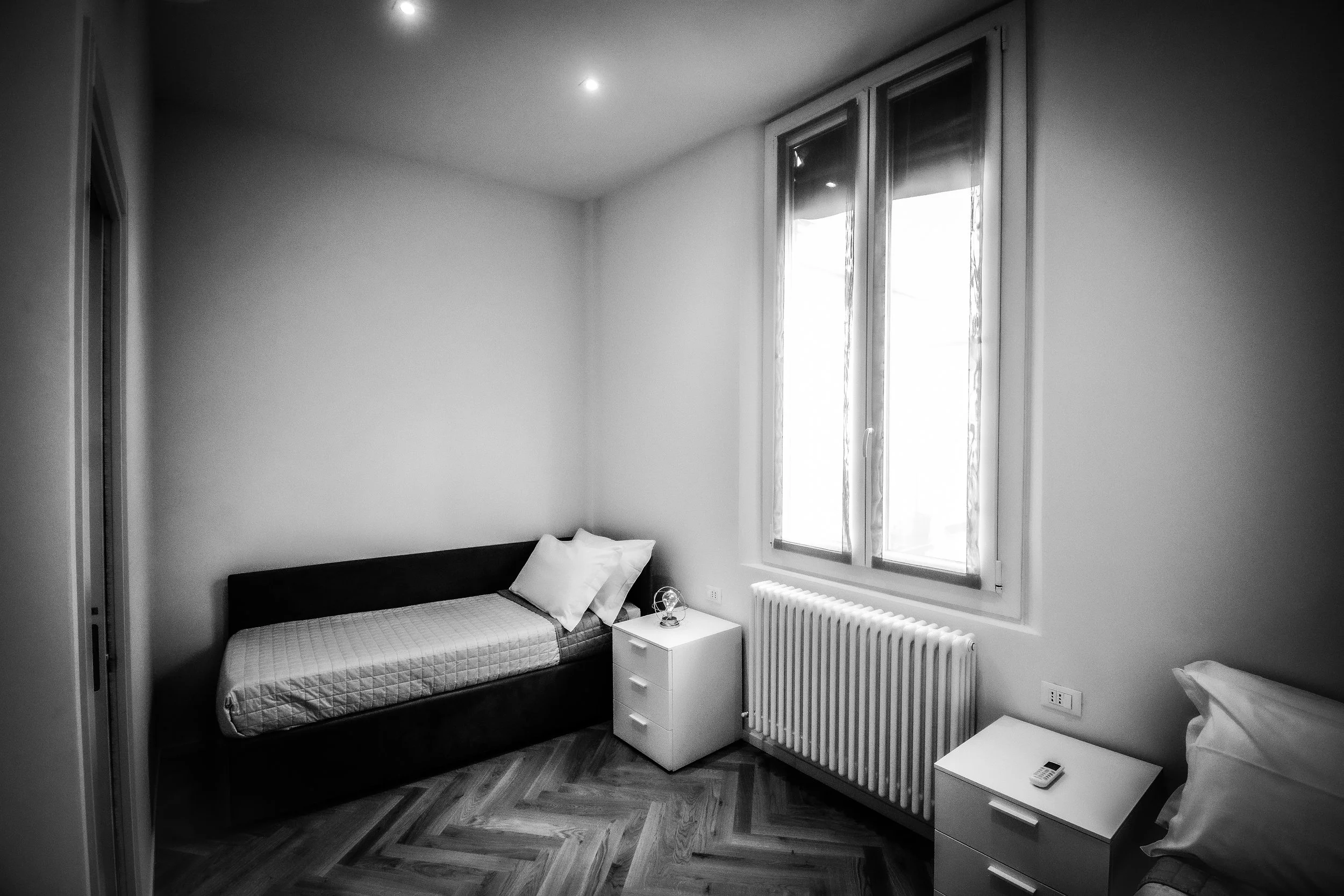 Camera con due letti singoli, finestra grande, due comodini e radiatore, senza decorazioni, in bianco e nero.