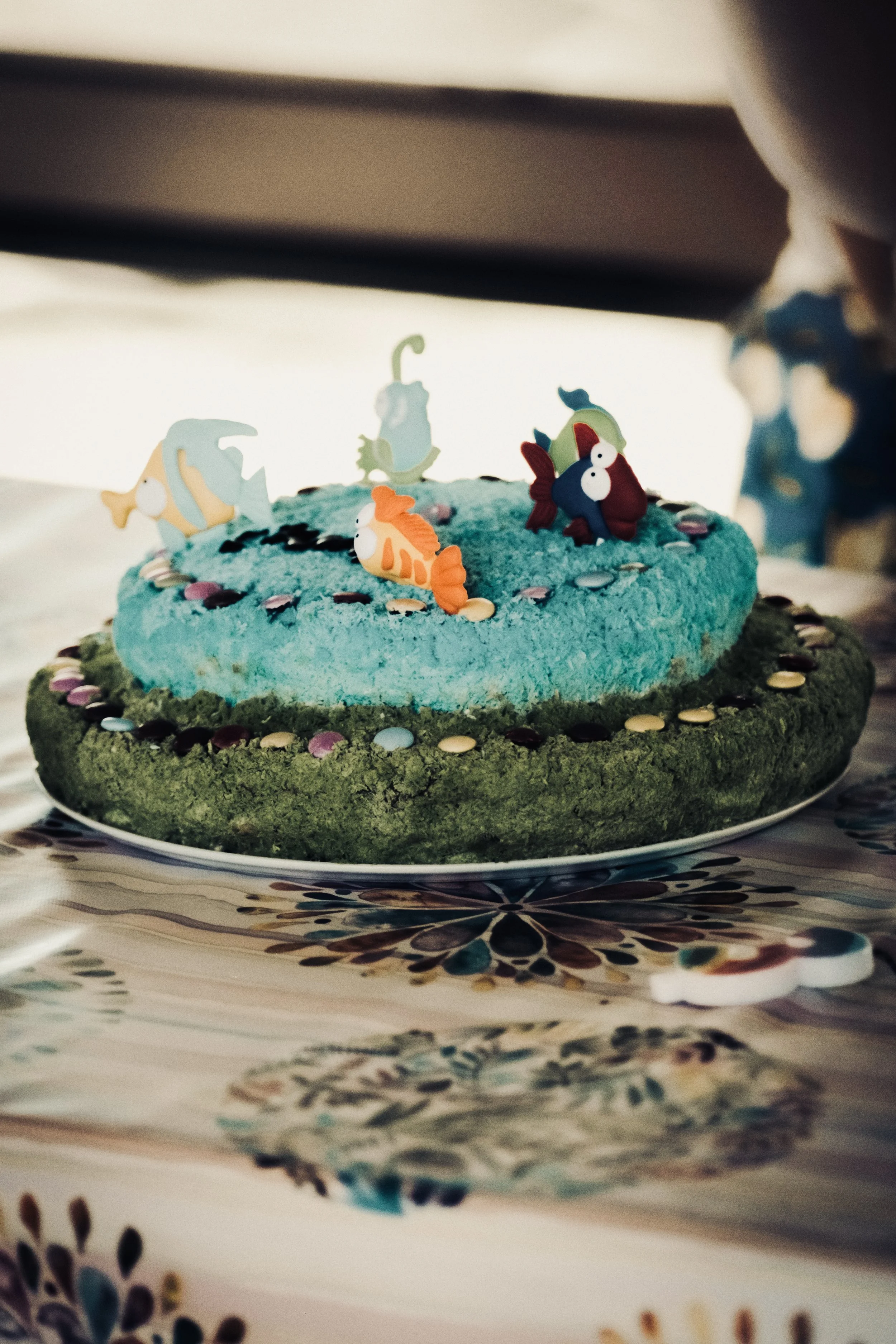 Torta decorata con elementi marini come pesci e alghe, con colore predominante azzurro e verde, posizionata su un piatto, su un tavolo con tovaglia decorata.