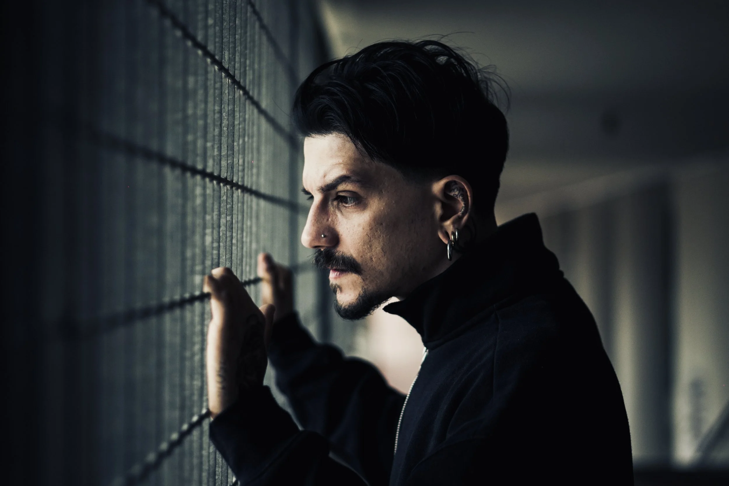 Uomo dai capelli corti neri, baffi e orecchini, guarda pensieroso attraverso una finestra con reagente alle pareti, indossa una giacca nera.