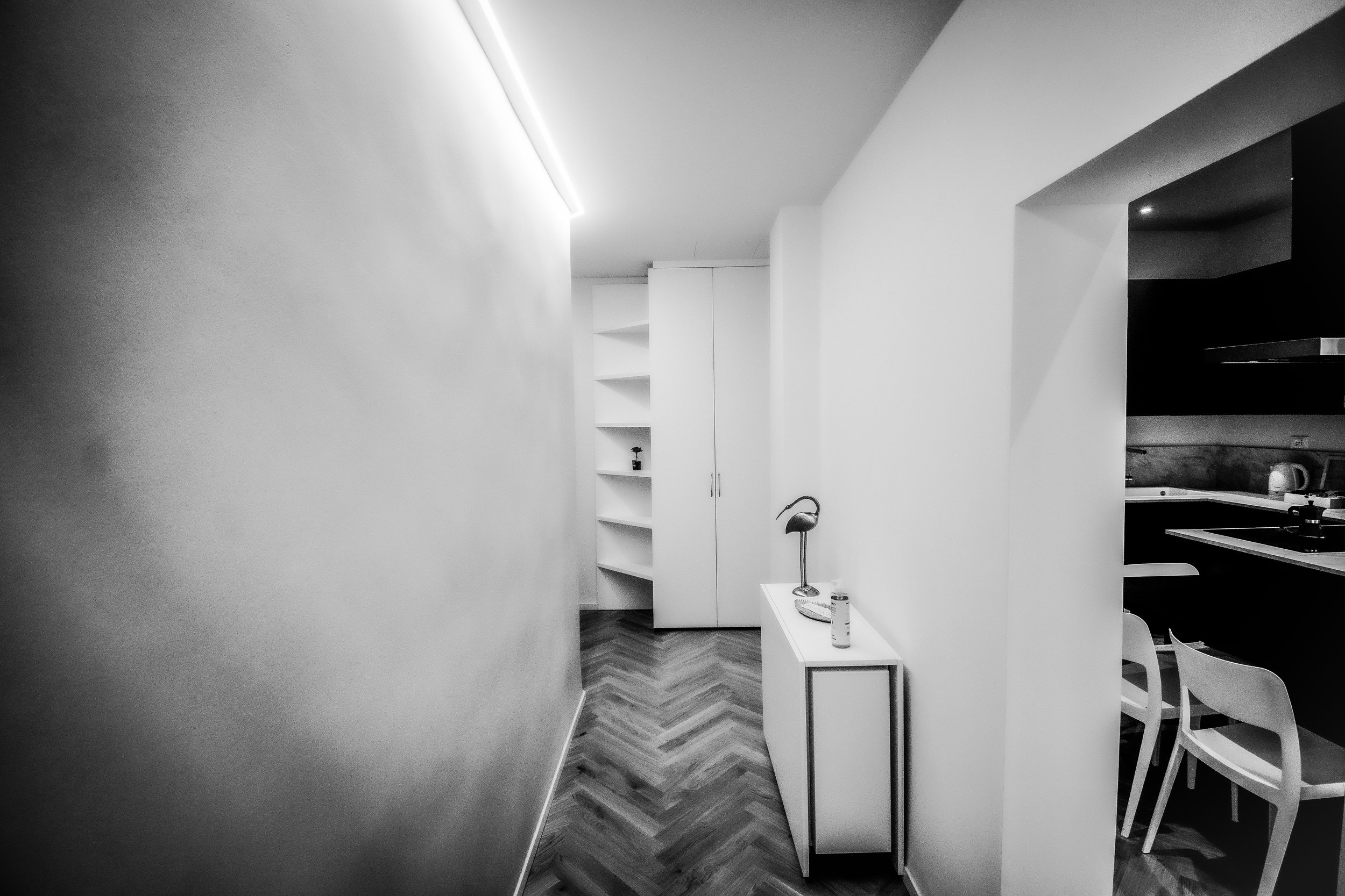 Interno di una casa con corridoio, pareti bianche, pavimento in parquet a spina di pesce, porta a battente, scaffale bianco con oggetti, cucina parzialmente visibile con sedie e tavolo.