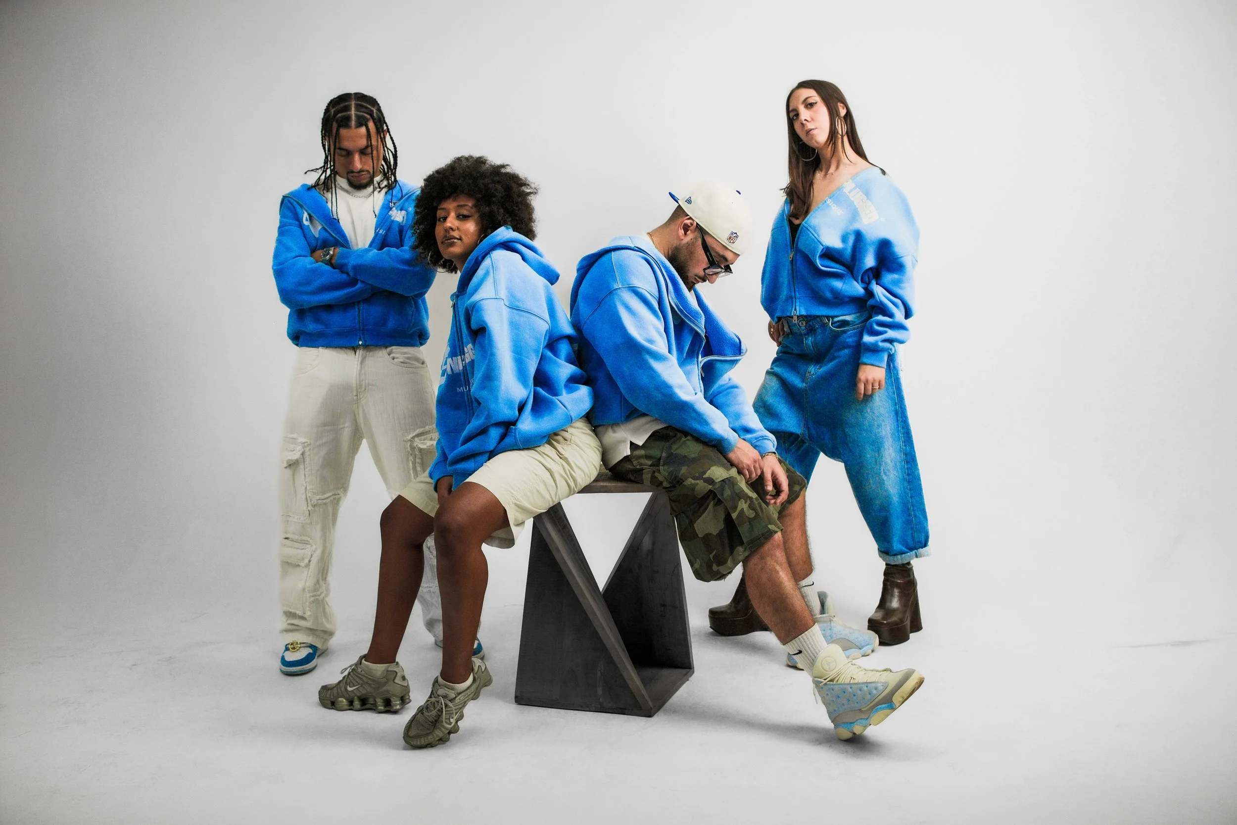 Cinque persone in abbigliamento casual posano in uno studio con sfondo bianco, alcune sedute e altre in piedi, indossano cappotti e sneakers di colore blu.
