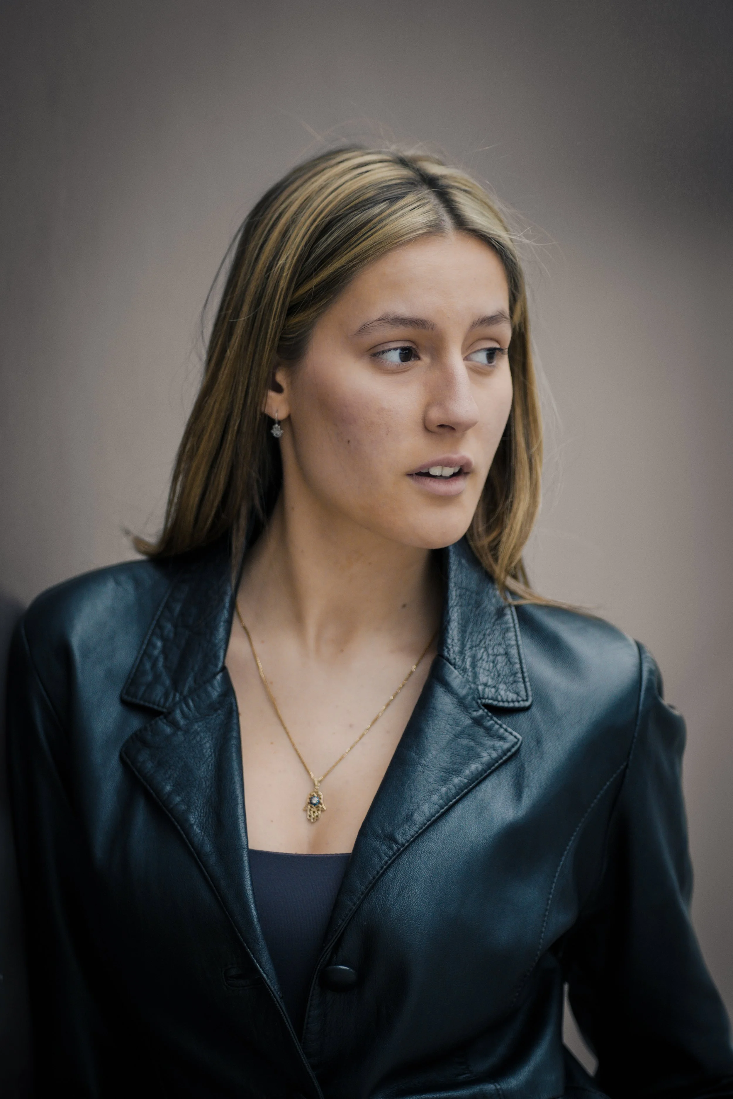 Una giovane donna con i capelli castani chiari e occhi azzurri, indossa una giacca in pelle blu scuro e una collana d'oro con un ciondolo. È rivolta di lato, con uno sguardo pensieroso.