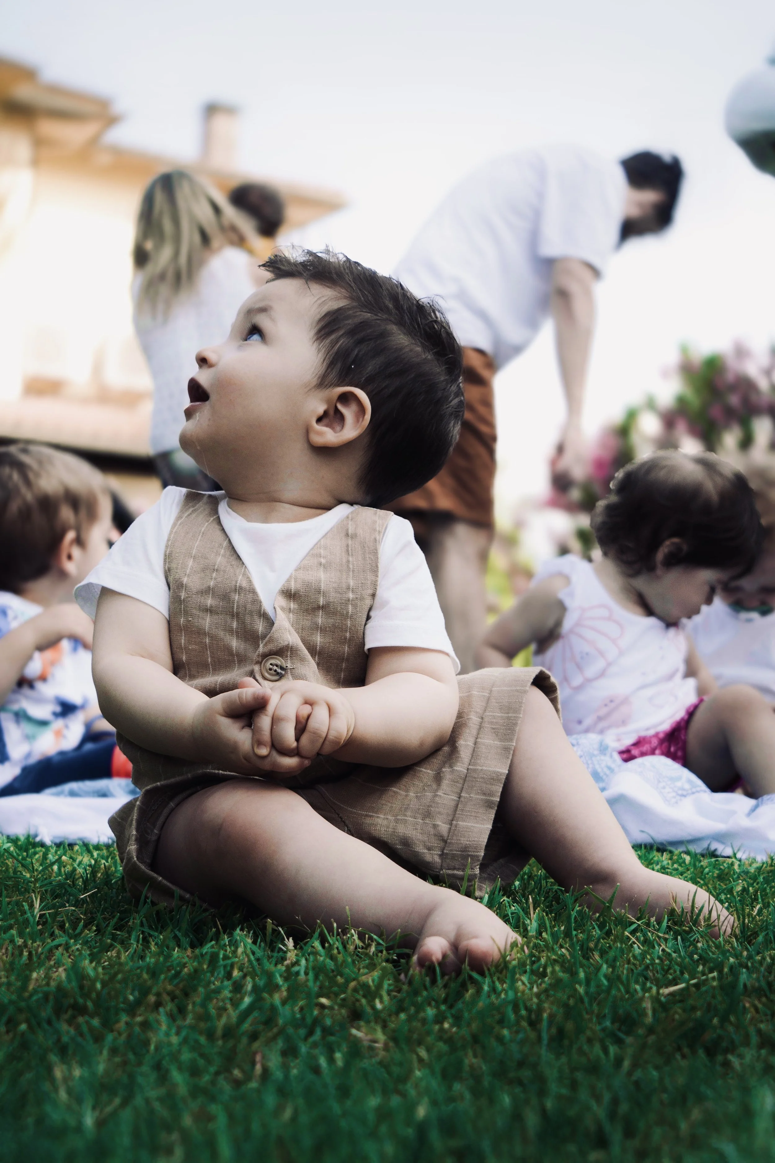 Un bambino seduto sull'erba, guarda verso l'alto con espressione stupita, circondato da altri bambini e adulti in un ambiente esterno.