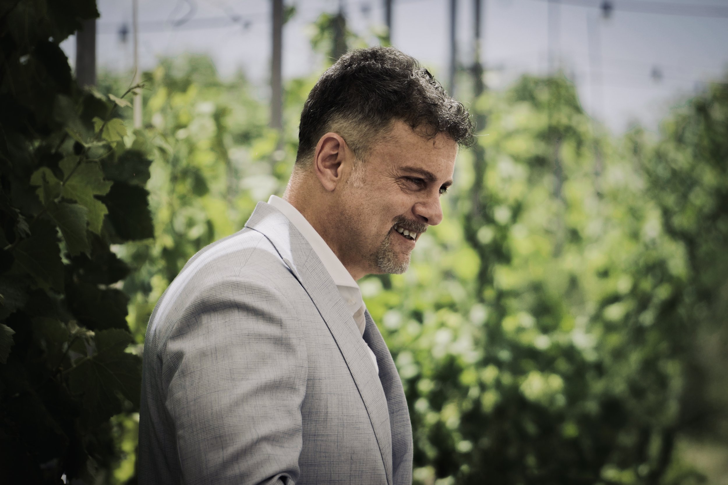 Uomo sorridente in abito chiaro che cammina tra piante verdi in una serra o giardino.