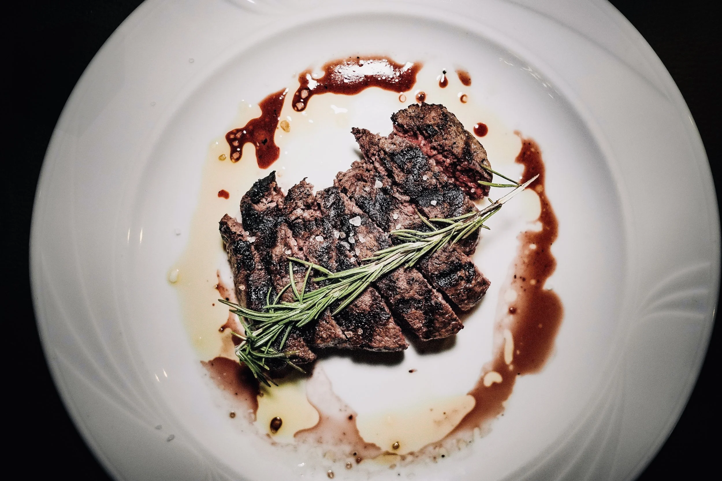 Bistecca di manzo con rosmarino sulla piastra, accompagnata da salse e olio, servita su un piatto bianco.