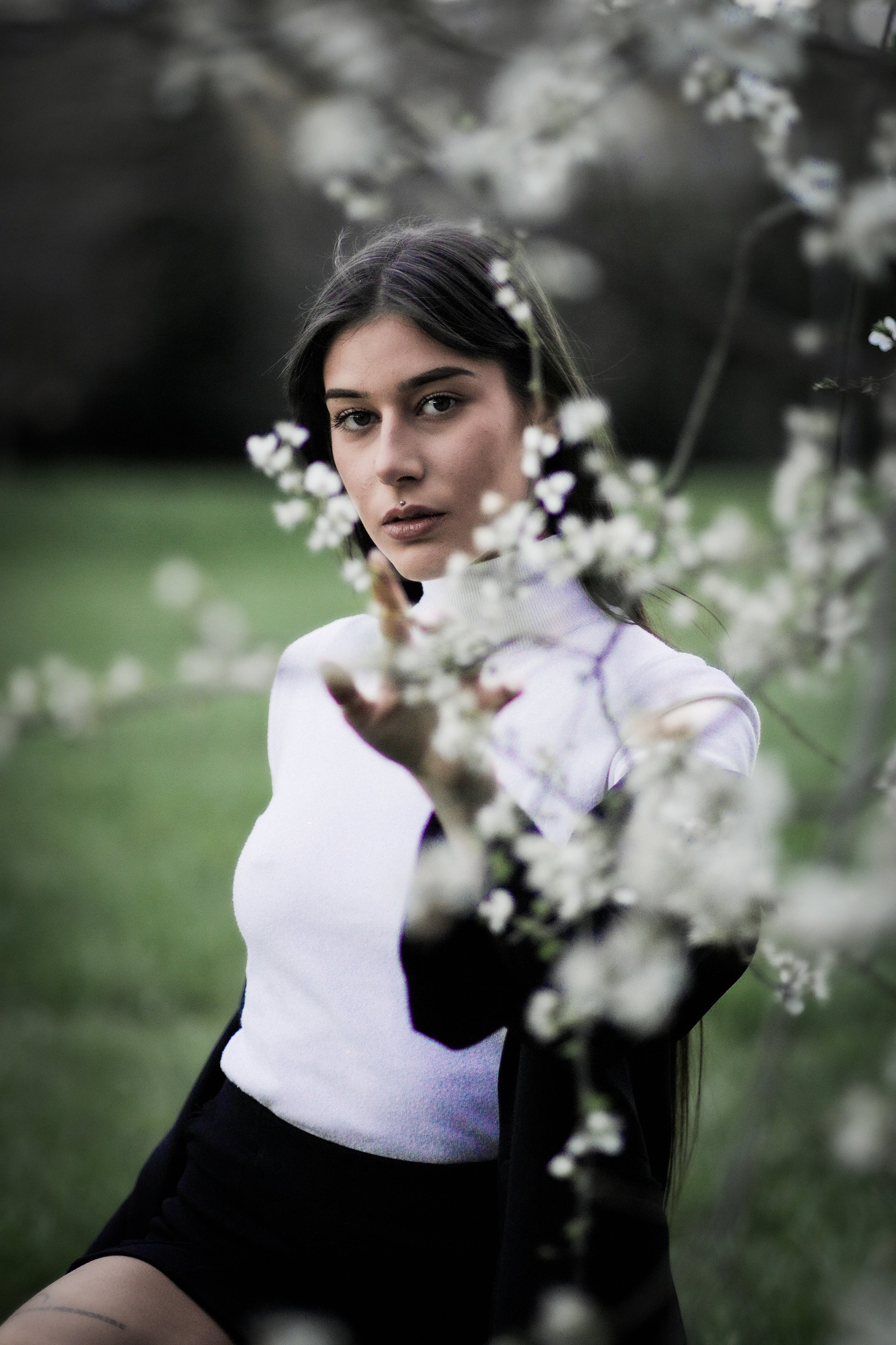 Ragazza vestita con top bianco e giacca nera che guarda verso la fotocamera attraverso rami di fiori bianchi.