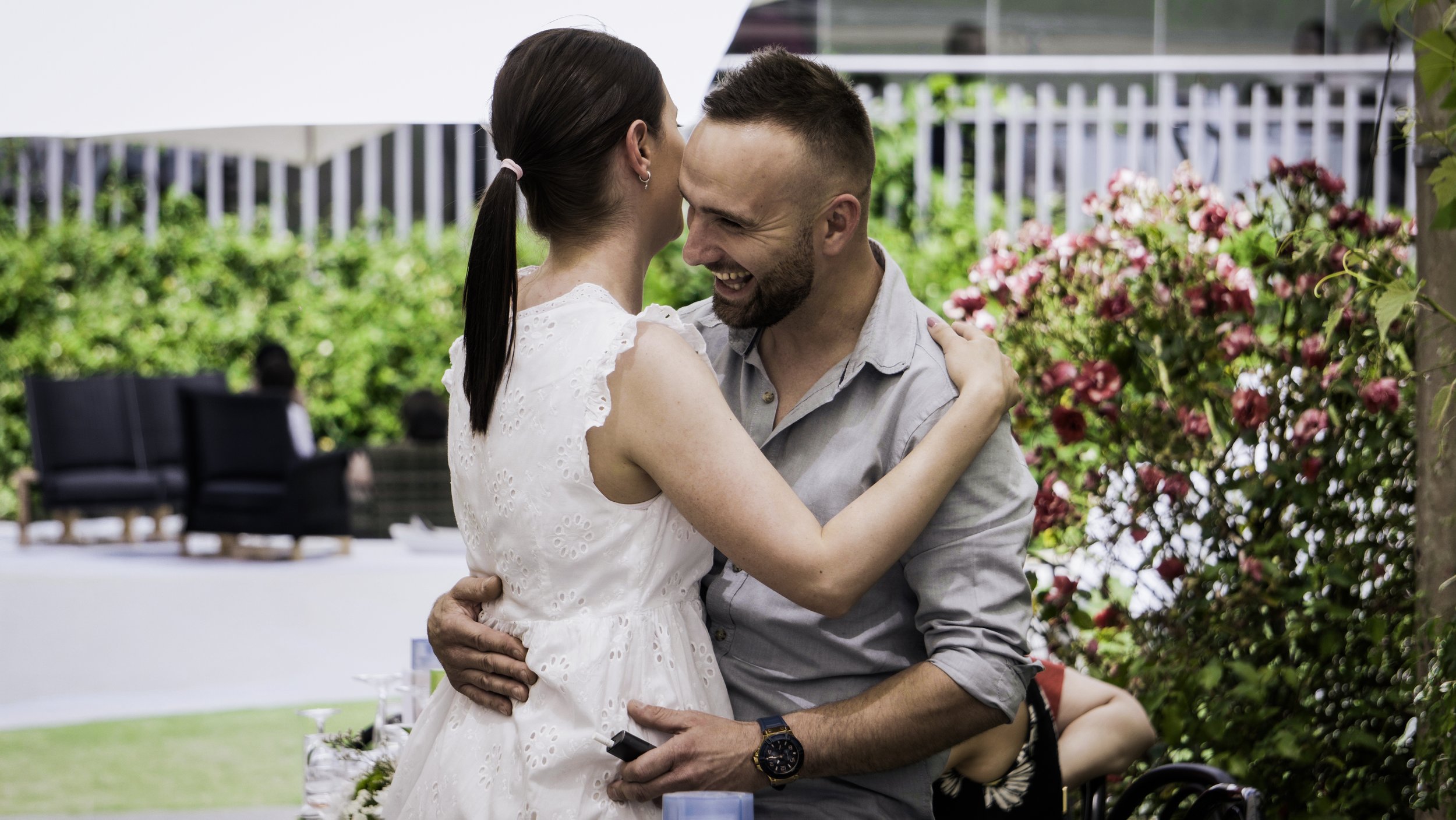 Una coppia abbracciata e felice in un giardino con fiori rosa, uno dei due con maglietta grigia, l'altra con vestito bianco.