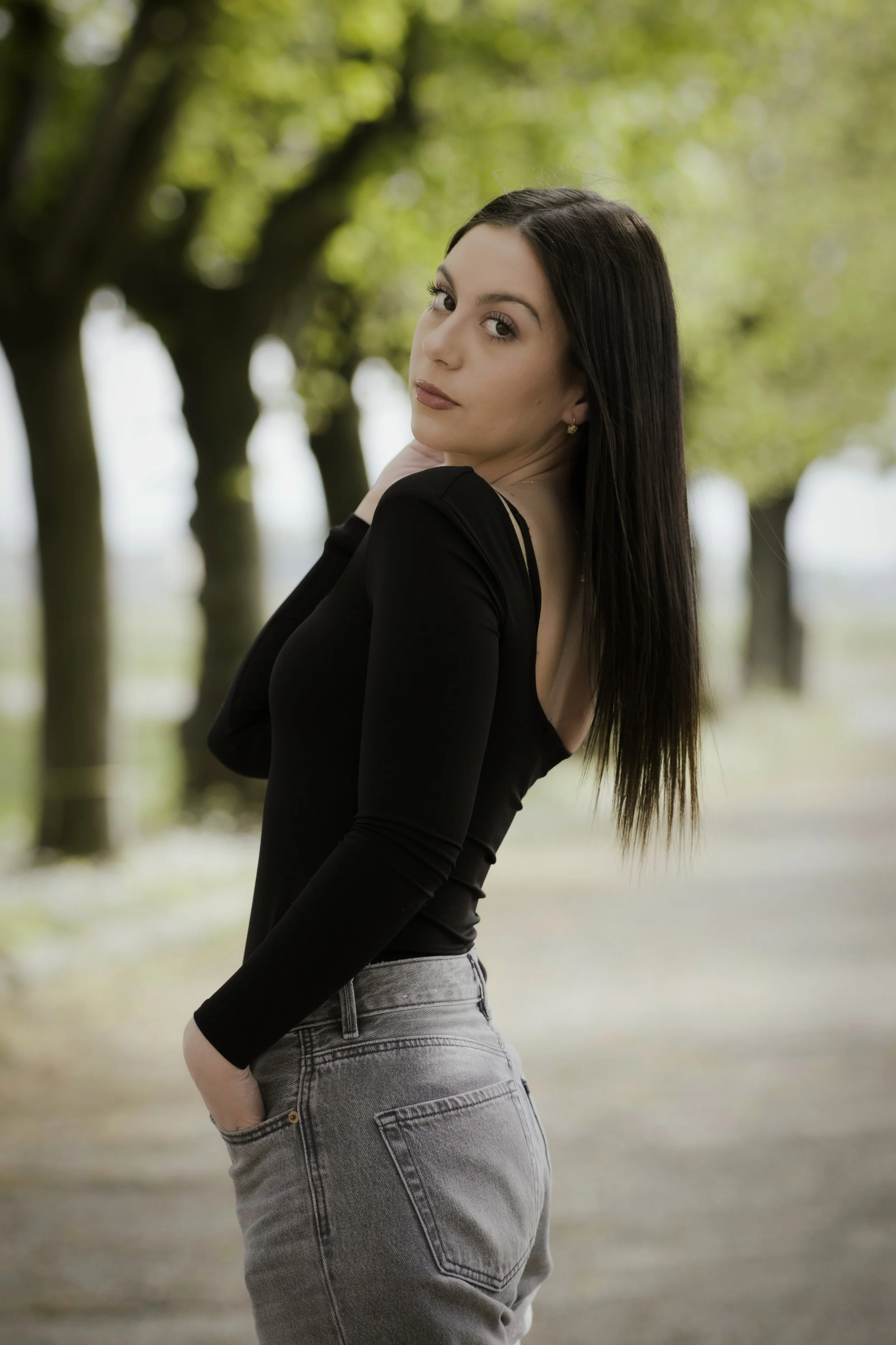 Ragazza con capelli lunghi, vestita con maglia nera e jeans grigi, che posa all'aperto tra gli alberi.
