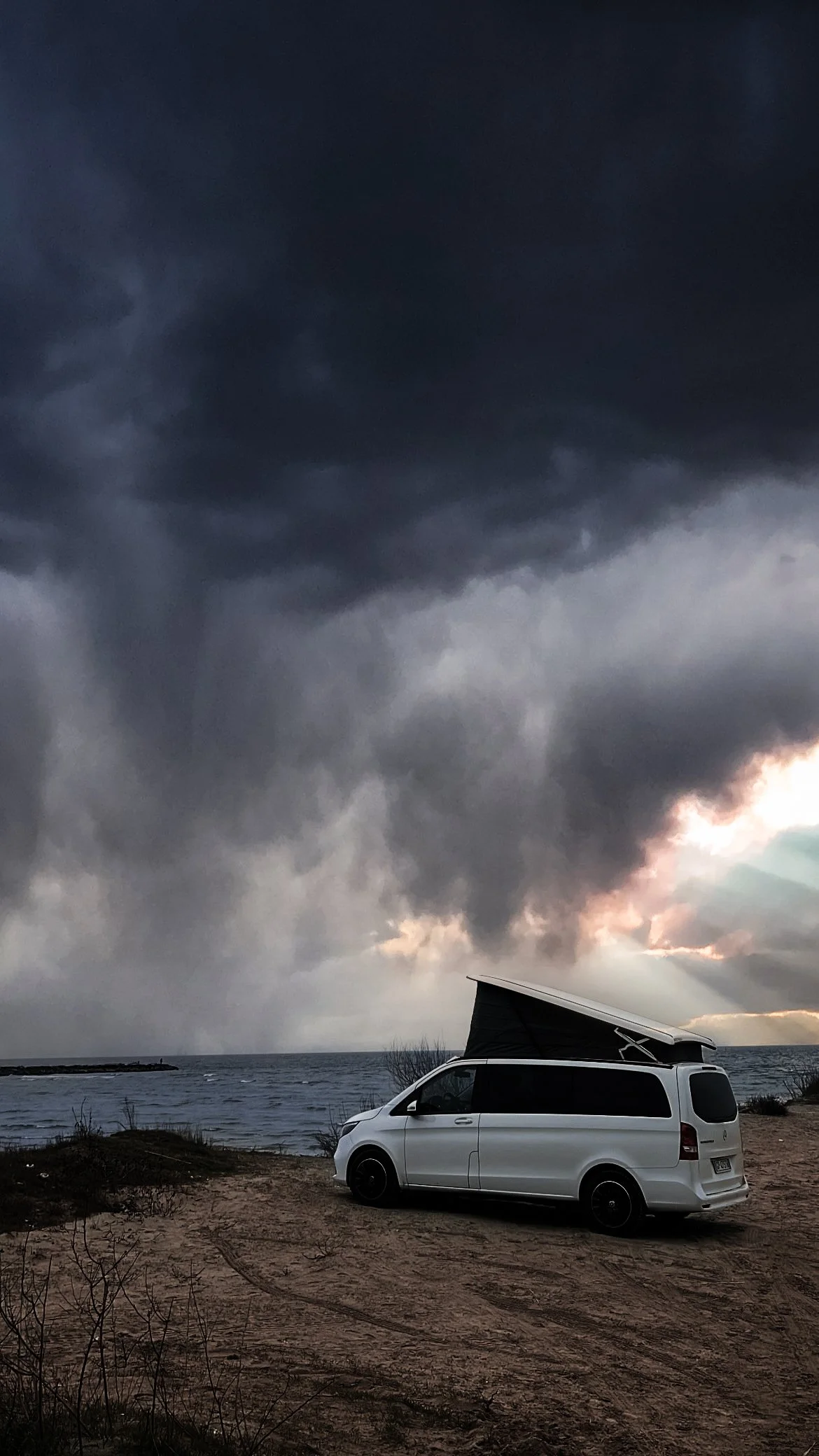 Un veicolo bianco con un tetto apribile parcheggiato su una spiaggia vicino al mare sotto un cielo scuro di temporale