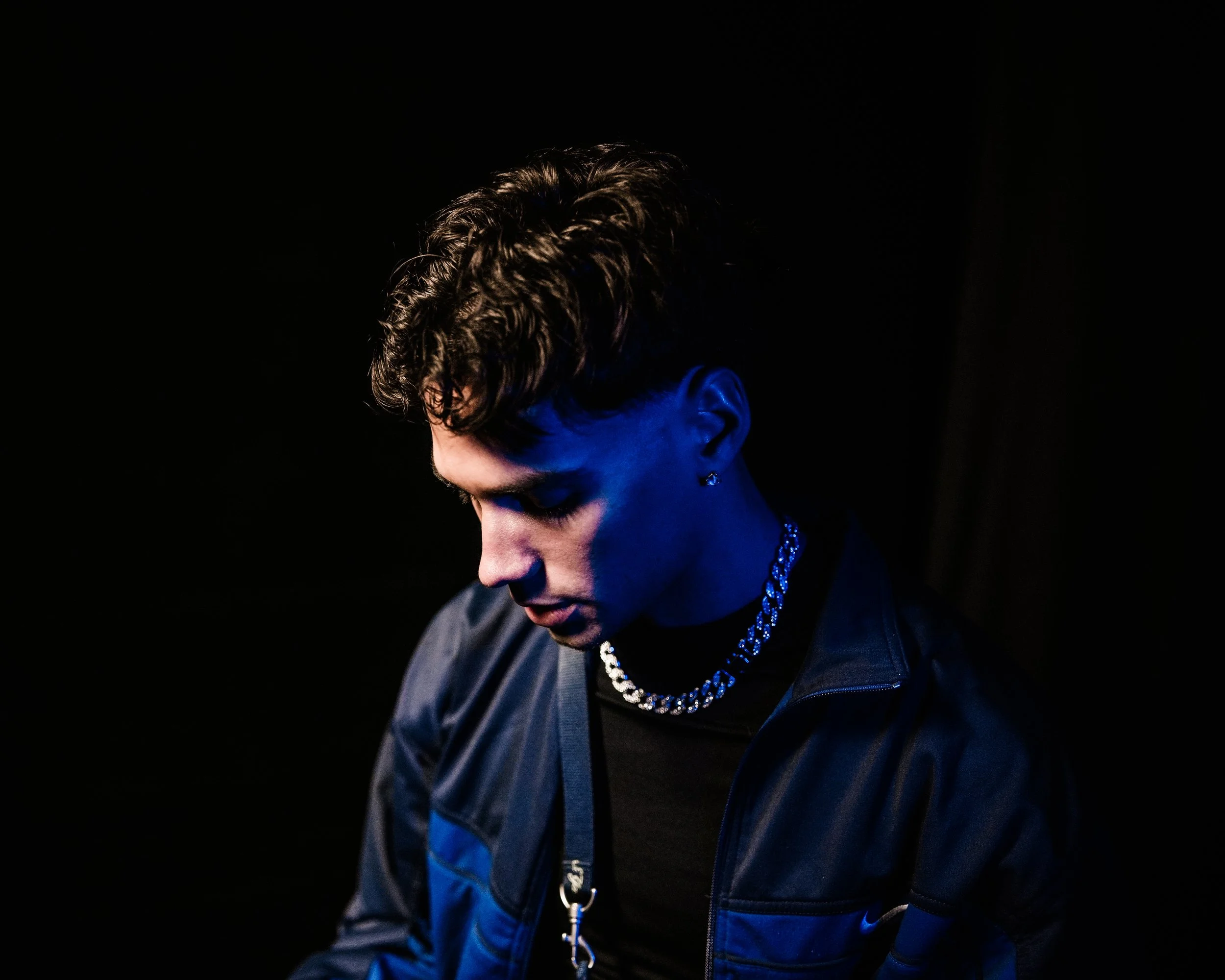 Ragazzo con capelli riccioli corti guarda verso il basso, indossa una catena argentata e una giacca scura, sfondo nero.