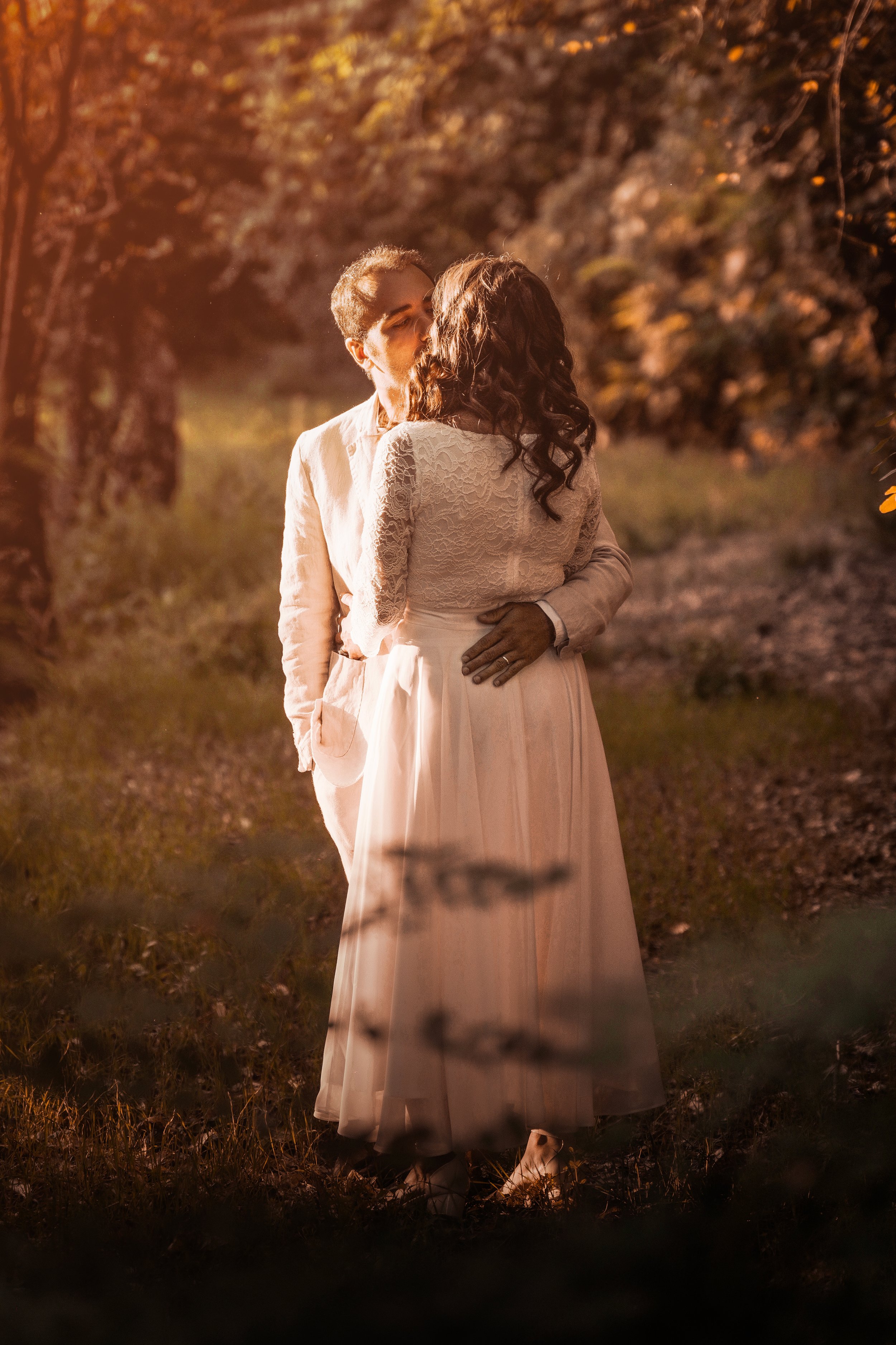 Una coppia di persone in abiti eleganti si baciano in un ambiente naturale con luci calde.