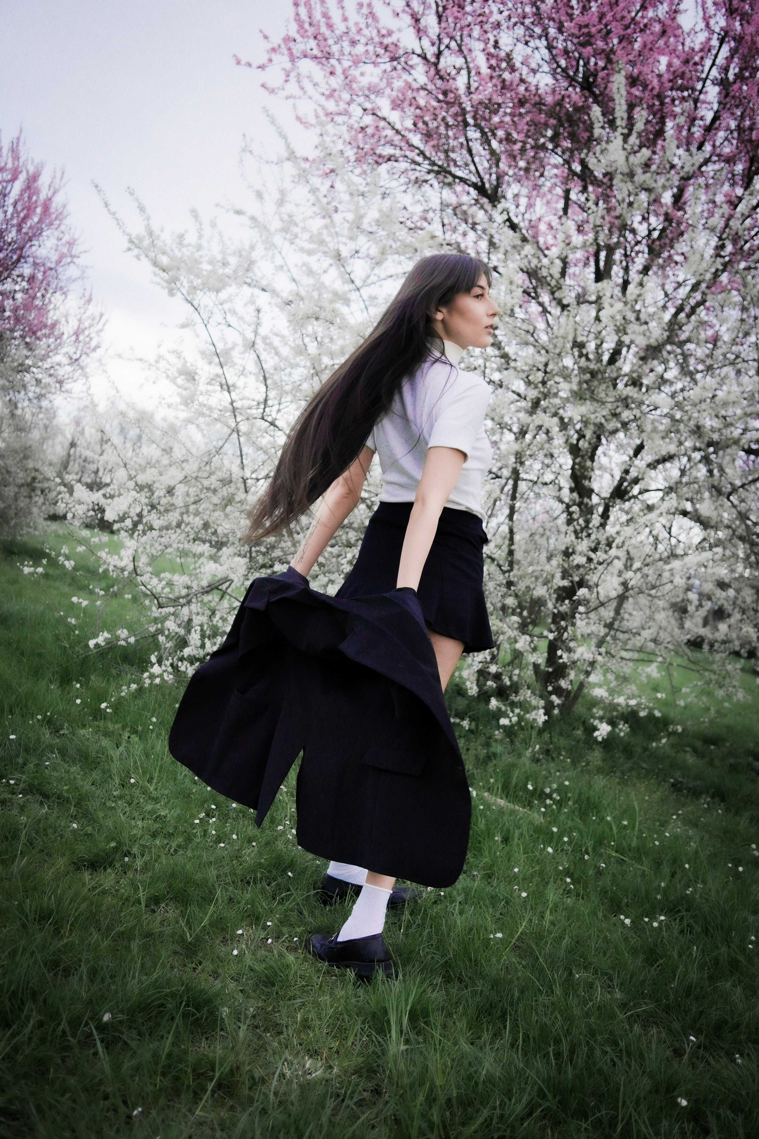 Una giovane donna con lunghi capelli scuri indossa un'uniforme scolastica e tiene un cappotto nero mentre si trova in un campo verde con alberi in fiore rosa e bianco sullo sfondo.