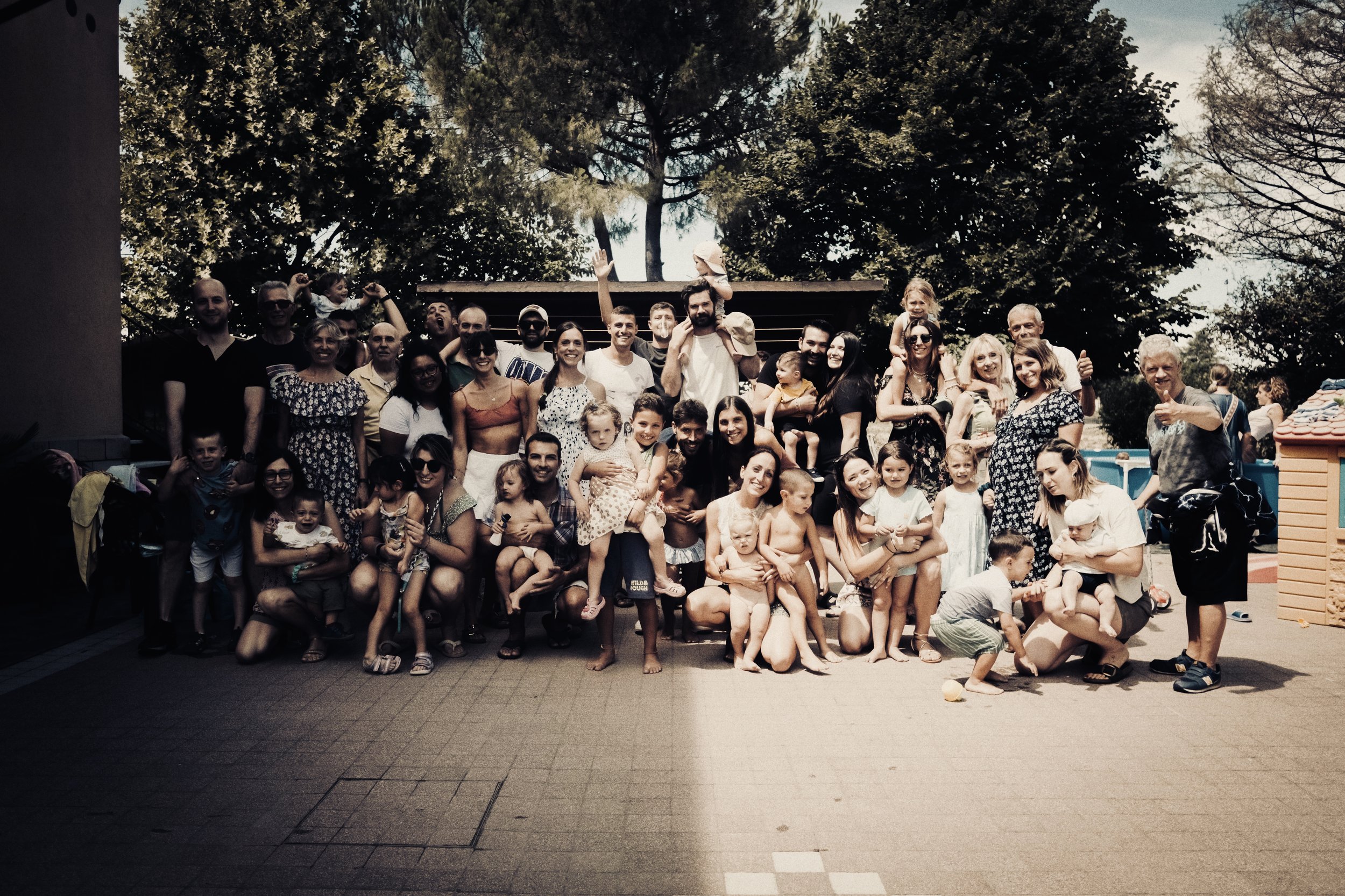 Un grande gruppo di persone di diverse età, tra adulti e bambini, che posano insieme all'aperto, con alcuni bambini sulle spalle e molti che sorridono e fanno gesti di approvazione.