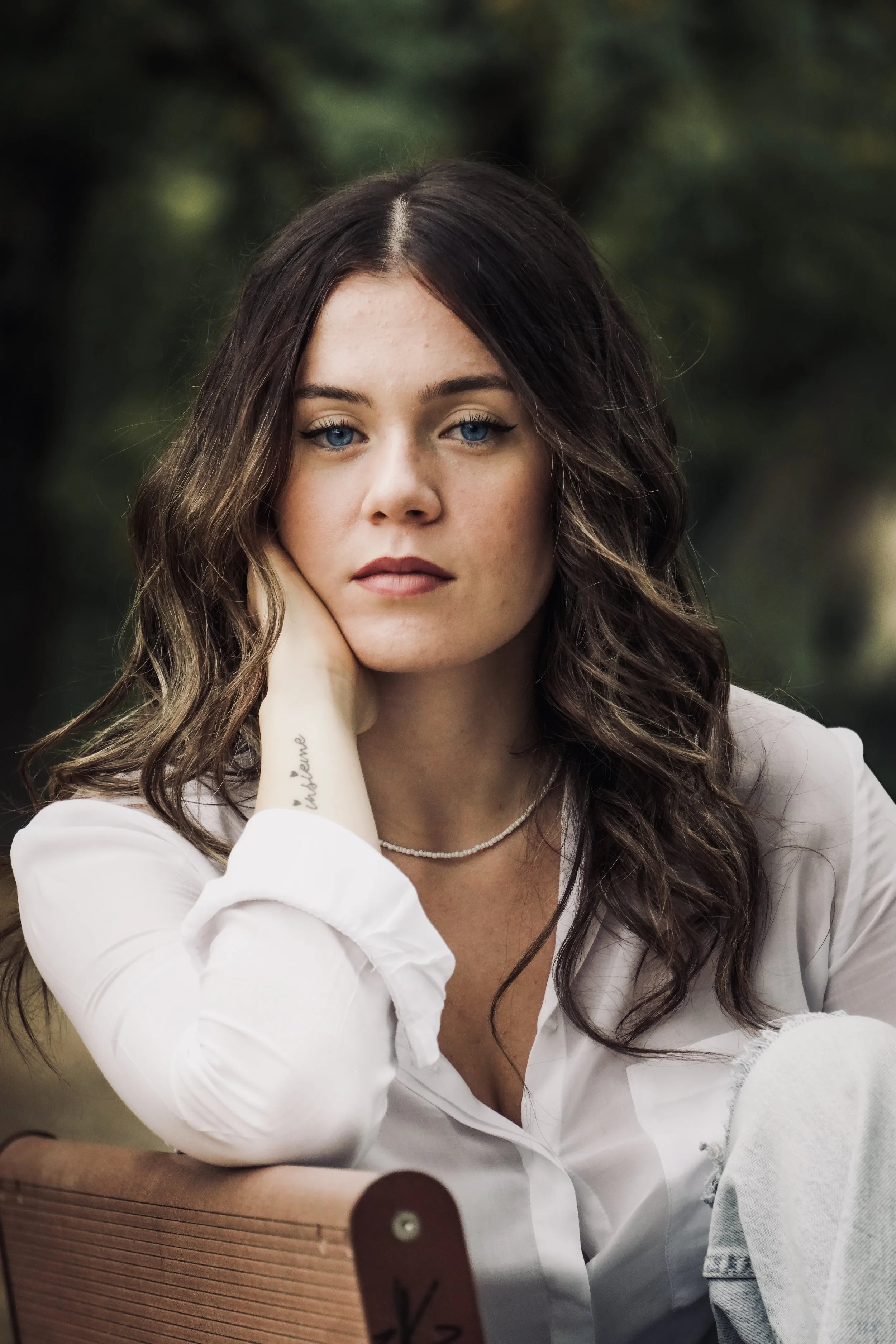 Ragazza con capelli castani ondulati, occhi blu, che si appoggia alla mano e guarda verso la fotocamera, con uno sfondo di natura piana e alberi.