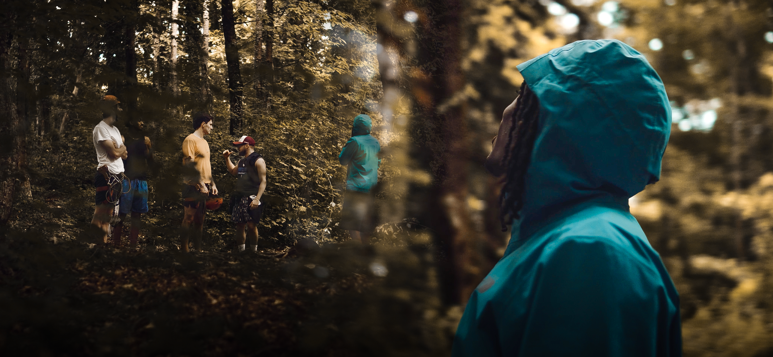 Un gruppo di persone con attrezzature da arrampicata in un bosco, e una persona in primo piano con cappuccio blu, che osserva il gruppo.