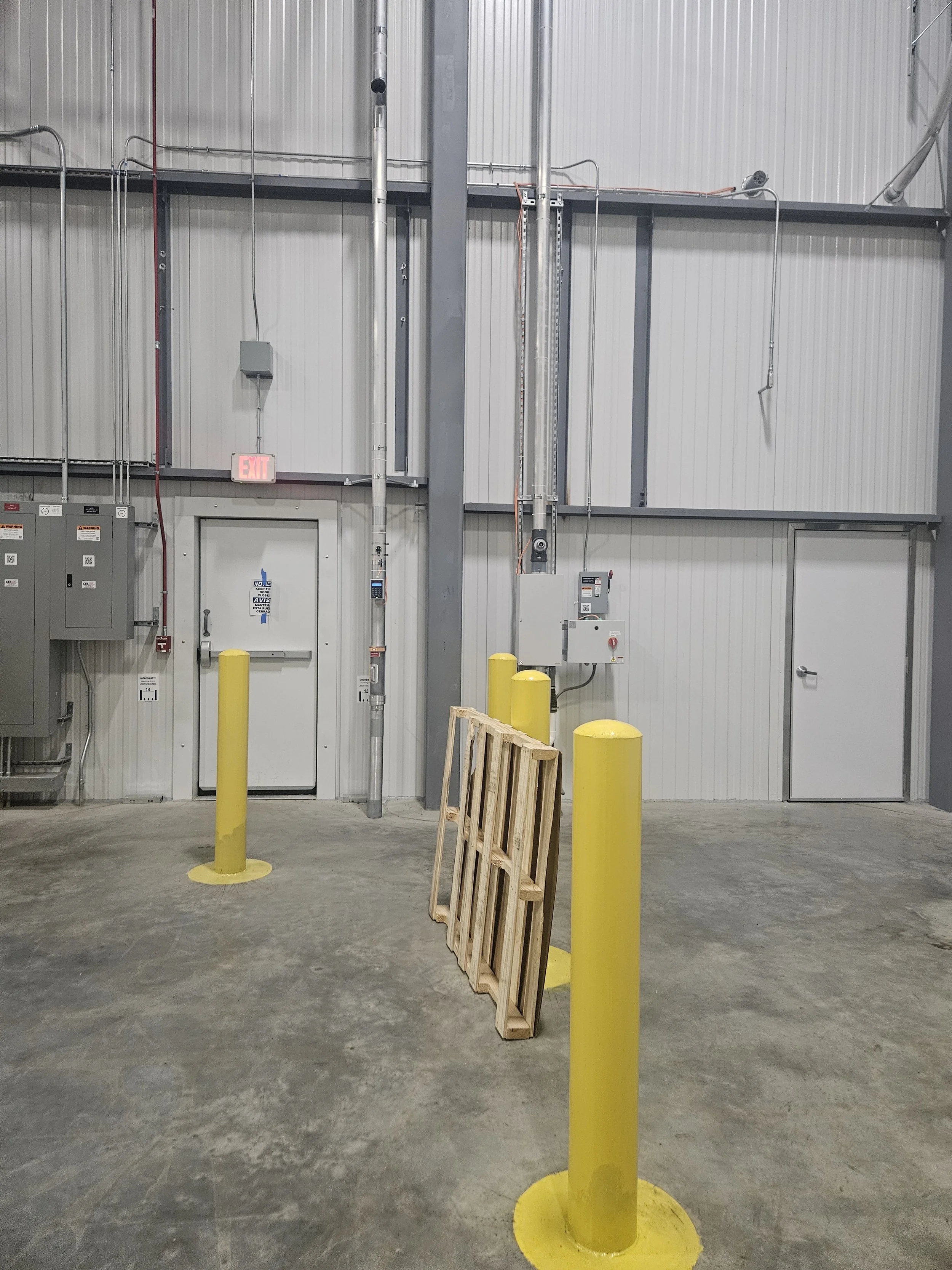 Industrial warehouse with gray metal walls, yellow safety bollards, a wooden pallet leaning against one bollard, and various electrical panels and piping on the wall.
