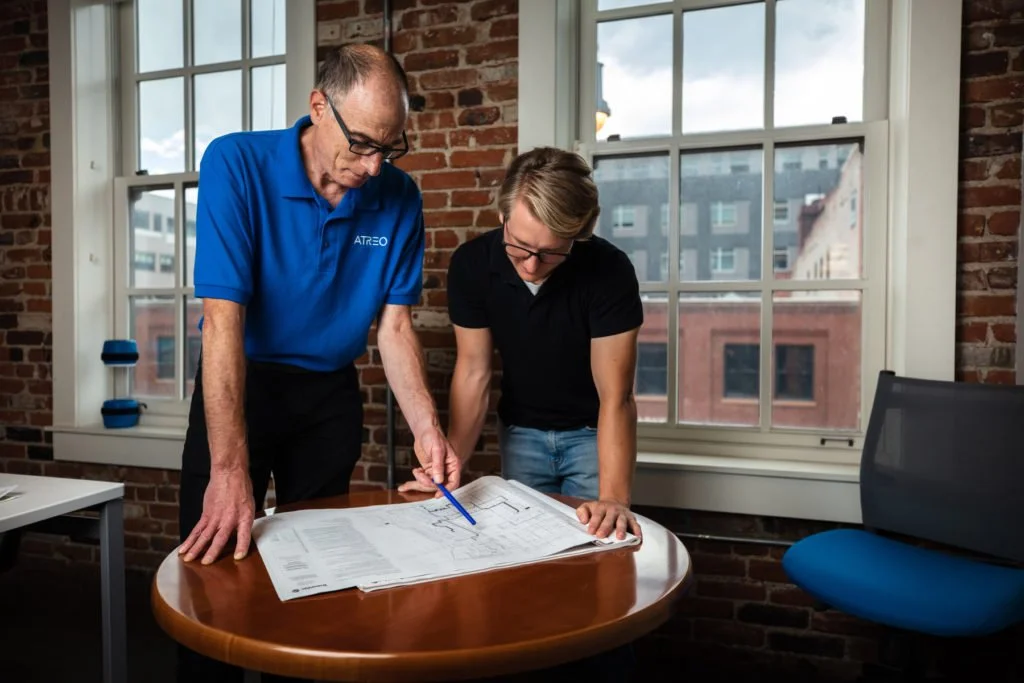 Two men examining architectural blueprints on a round table in an office with brick walls and large windows.