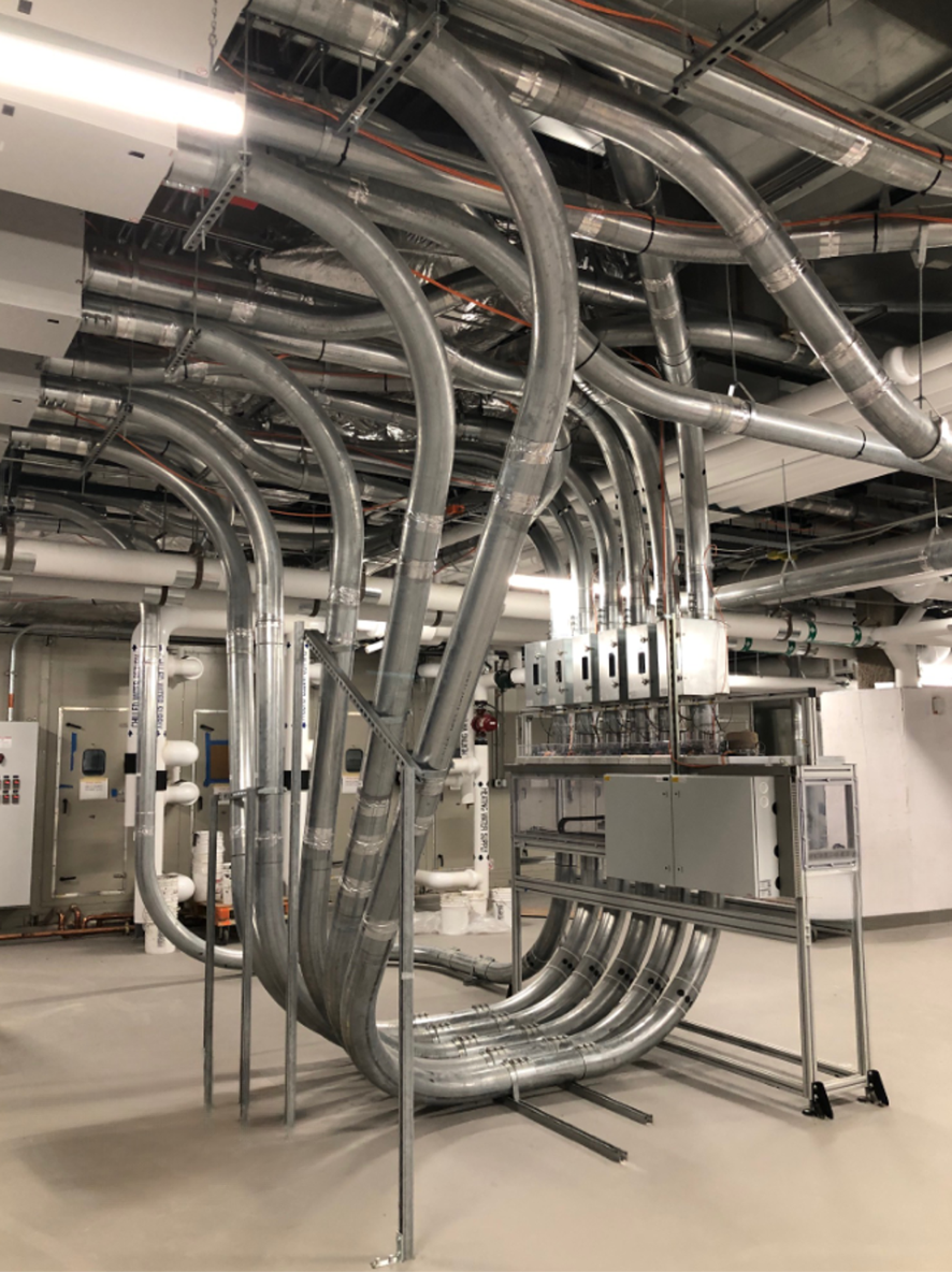 Interior of an industrial facility with numerous large metal pipes and conduits arranged overhead and on the floor, along with various electrical panels and equipment.