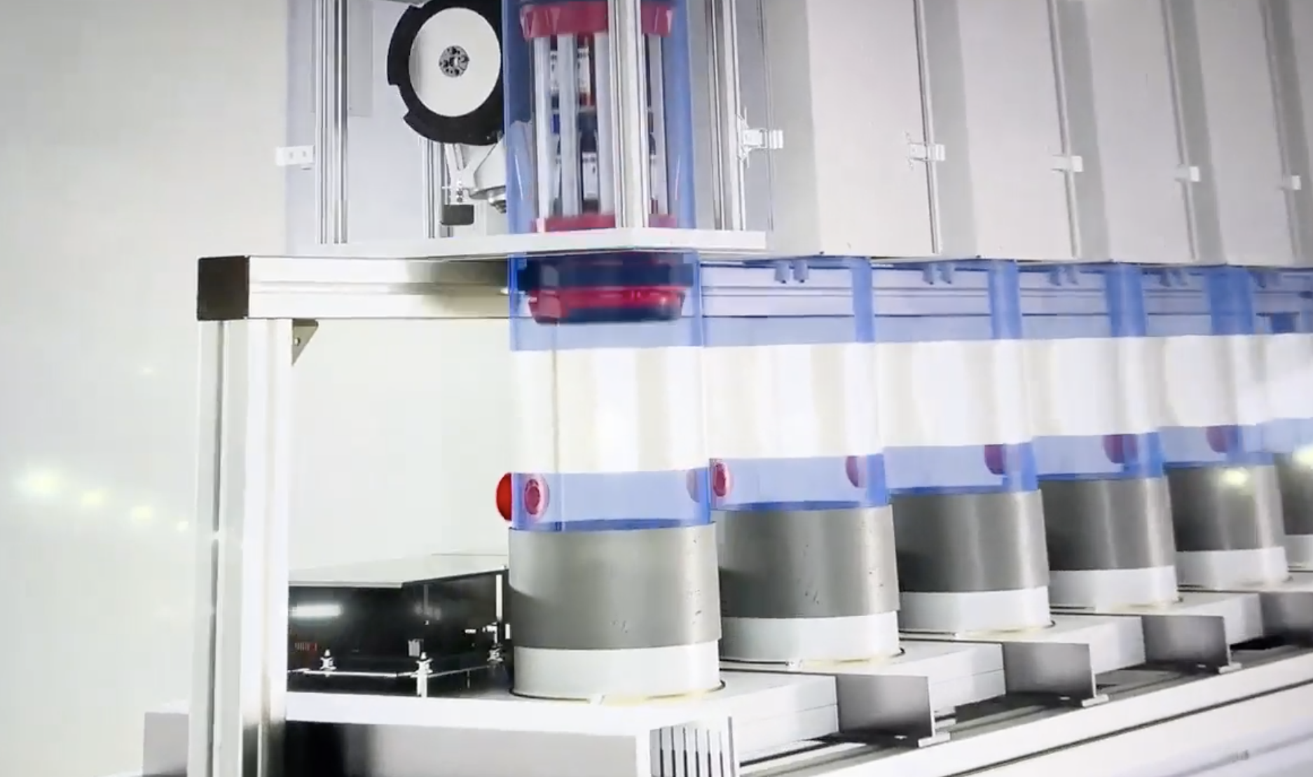 Automated manufacturing or laboratory equipment with robotic arms, conveyor, and vertical stacks of containers or test tubes in a clean, industrial setting.