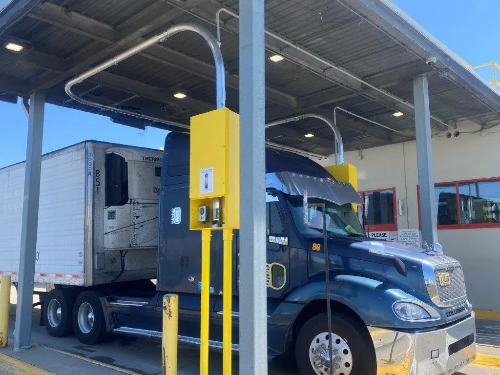 A semi-truck parked at a loading dock under a metal canopy, with a refrigerated trailer attached, yellow safety bollards, and a sign that says 'Please'.