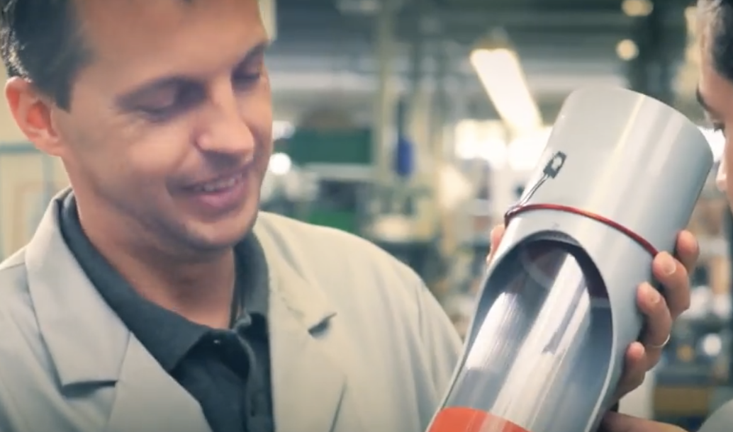 A man in a lab coat smiling and holding a long metallic tube or pipe in a workshop or industrial setting.