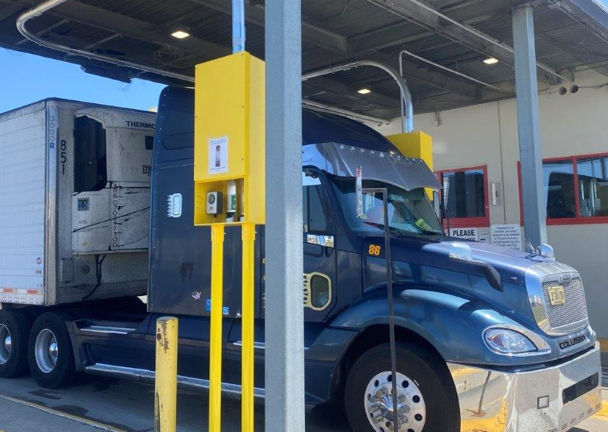 A semi-truck parked at a fueling station, with the truck's trailer attached and a yellow fuel pump nozzle inserted into the truck's fuel tank.