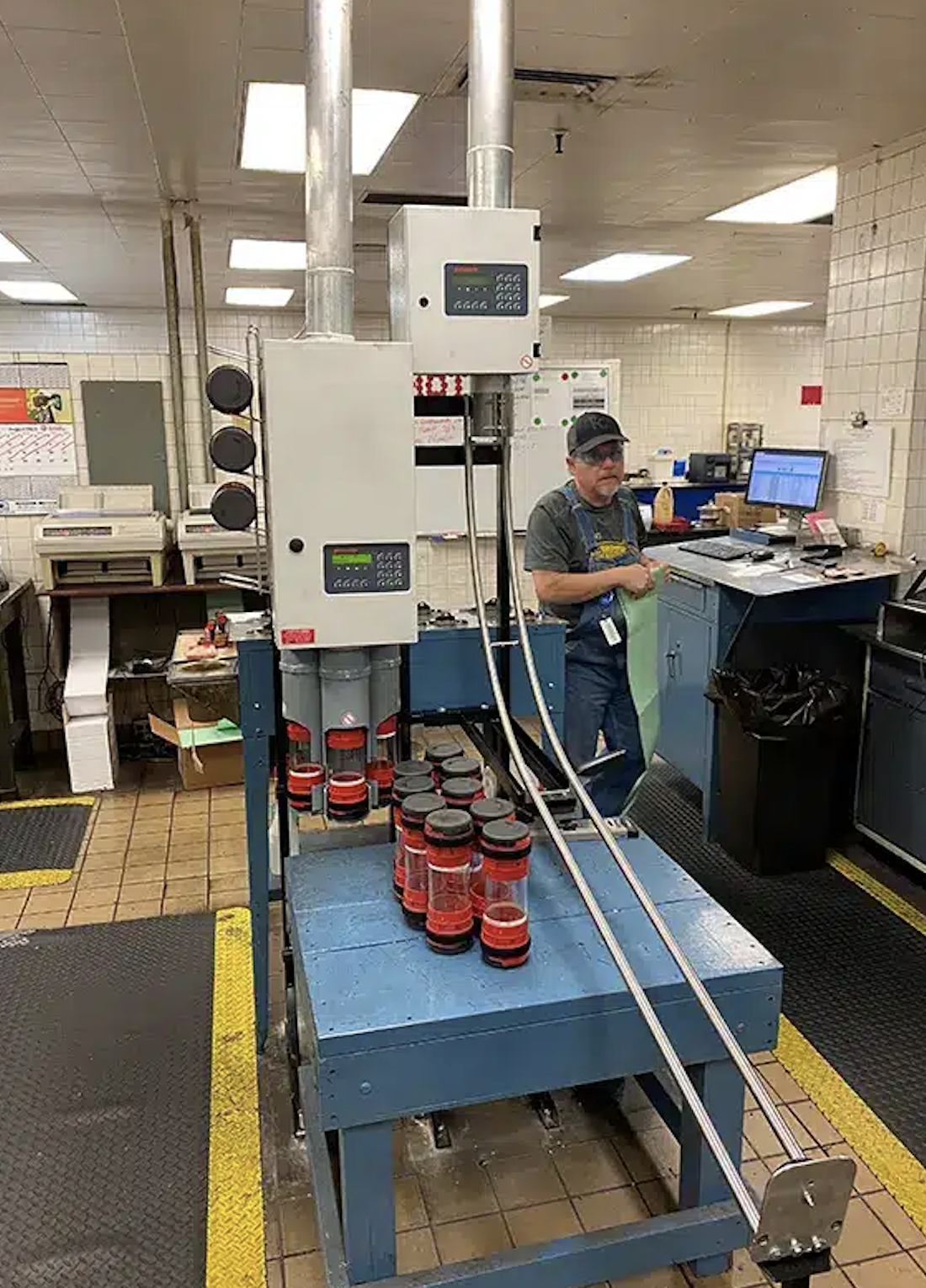 A man wearing a baseball cap and apron stands next to a machine in a commercial kitchen or factory setting, with jars lined up on a conveyor belt.
