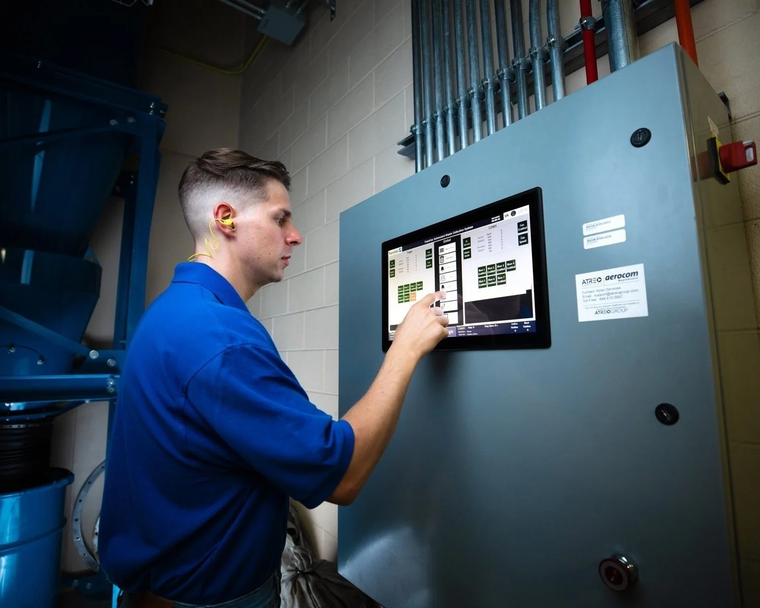 A man in blue work clothes using a touchscreen control panel in an industrial setting, with pipes and electrical wiring visible in the background.