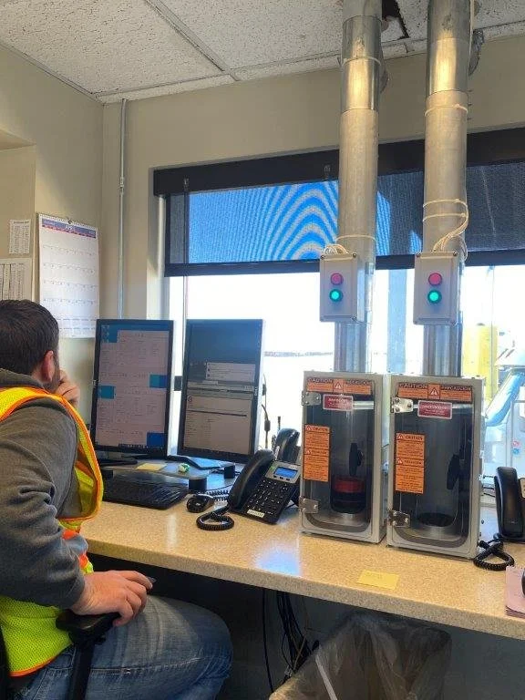 Office workstation with two monitors, a telephone, two safes with warning labels, and two electrical boxes above the safes, with man wearing a safety vest working at the desk.