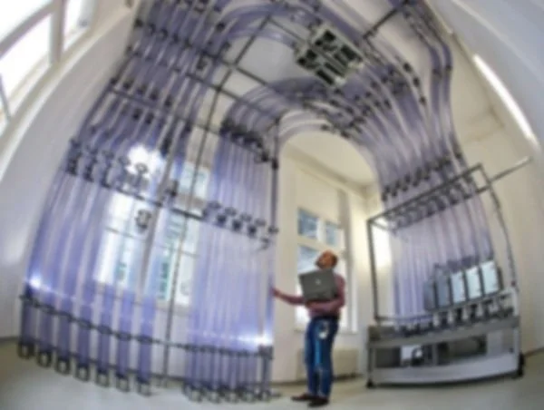 A person standing in a hospital or medical facility beside a large, complex medical equipment setup with various cables and tubes, possibly a life support or imaging system.