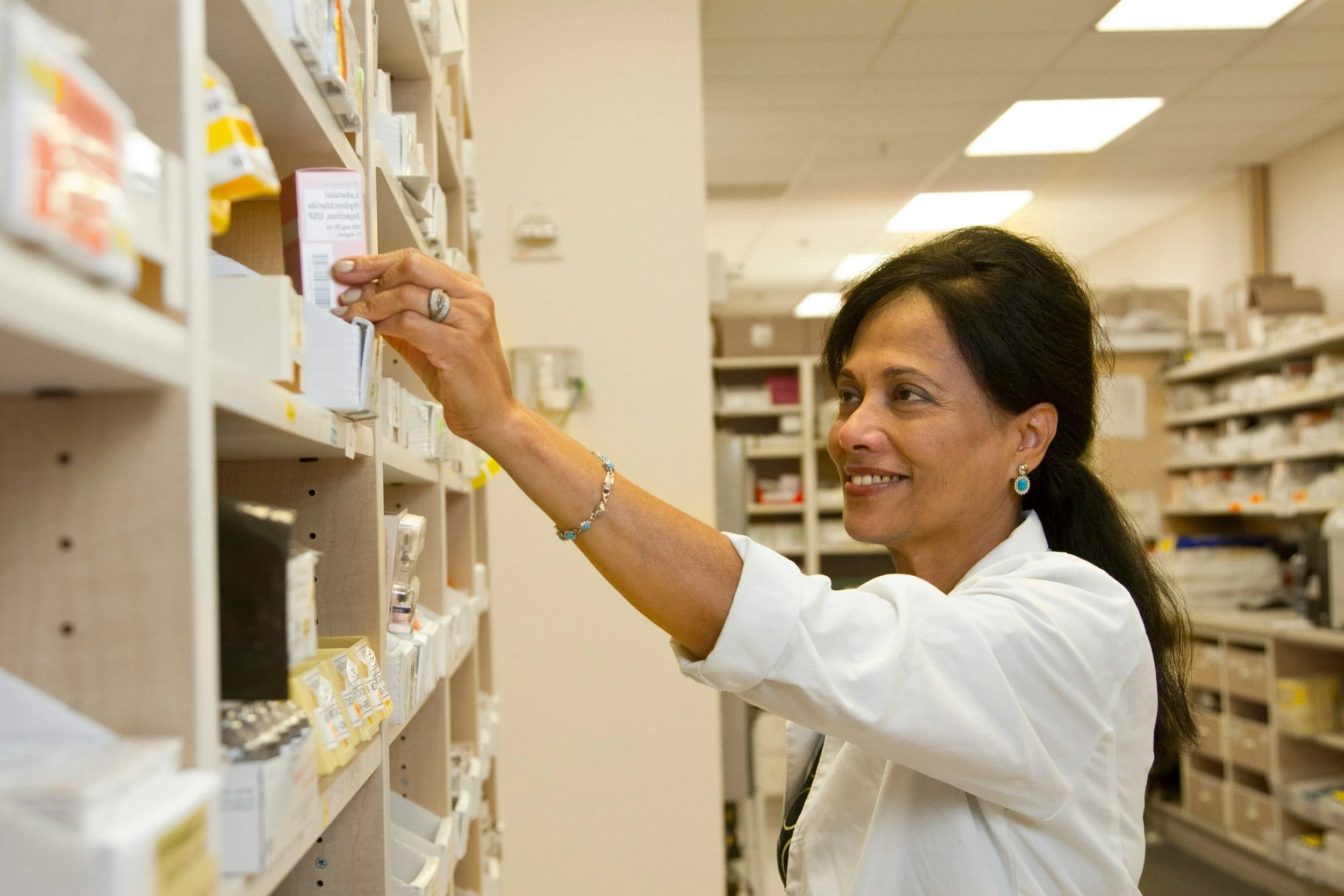 A woman in a white coat selecting medication from pharmacy shelves.