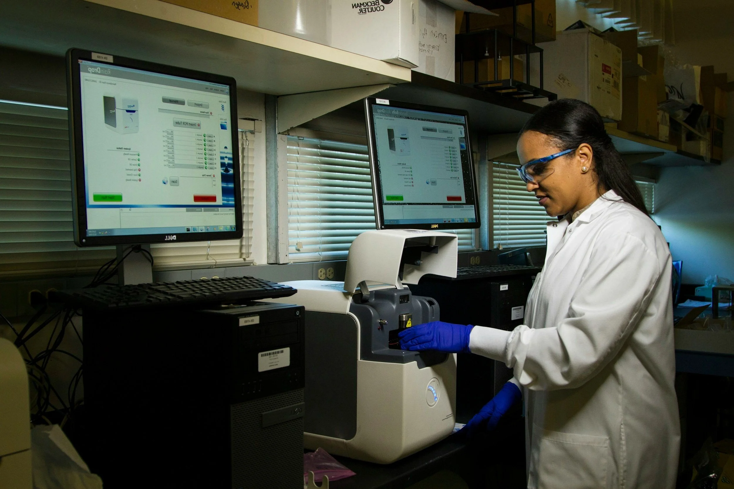 A scientist in protective glasses and gloves operating a laboratory instrument with multiple computer screens displaying data.