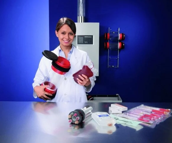 A female scientist in a white lab coat holding laboratory equipment and supplies in a lab with blue walls.