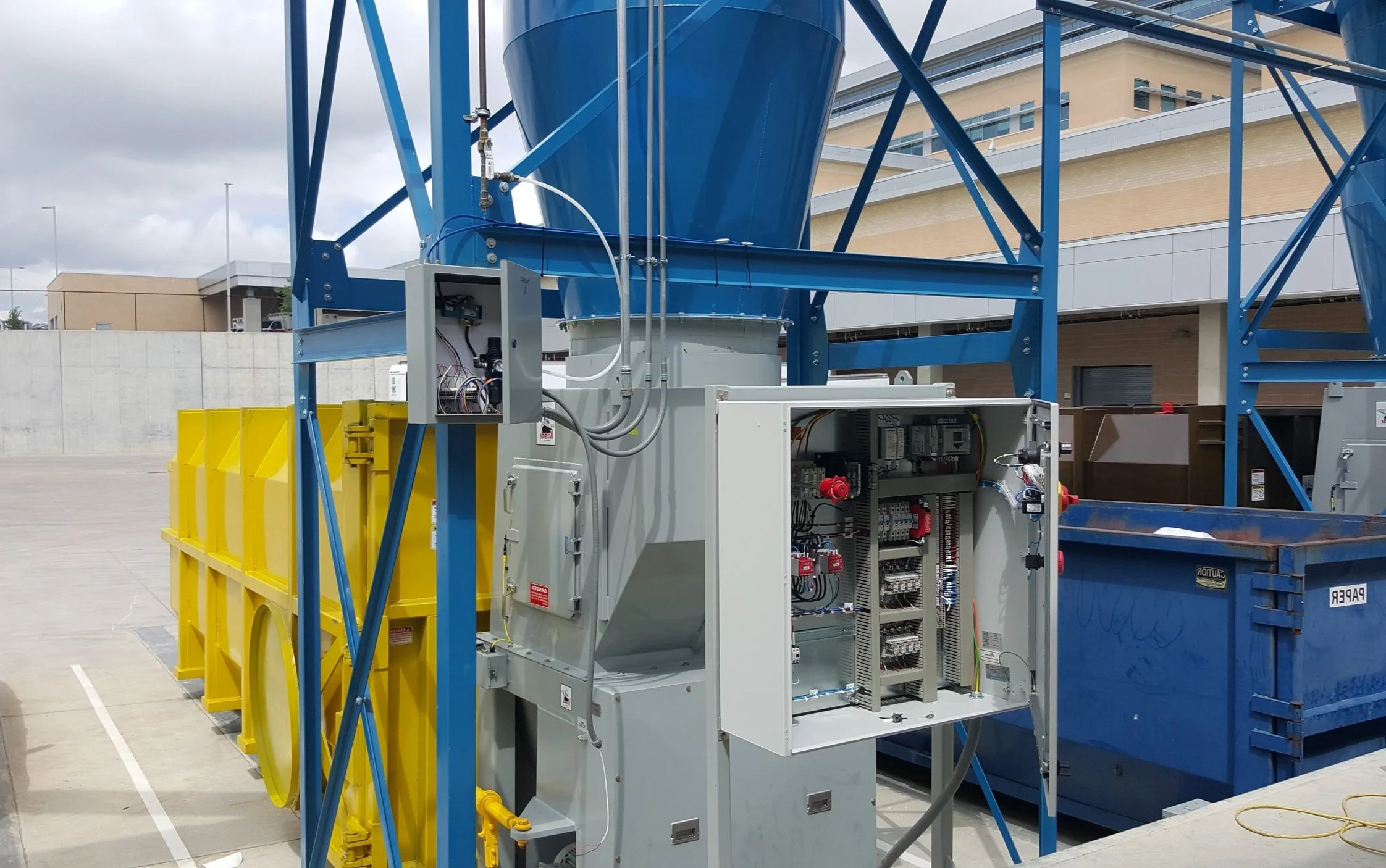 Electrical and control panels installed on a blue industrial structure with large yellow bins nearby, set in an outdoor parking lot with a building in the background.