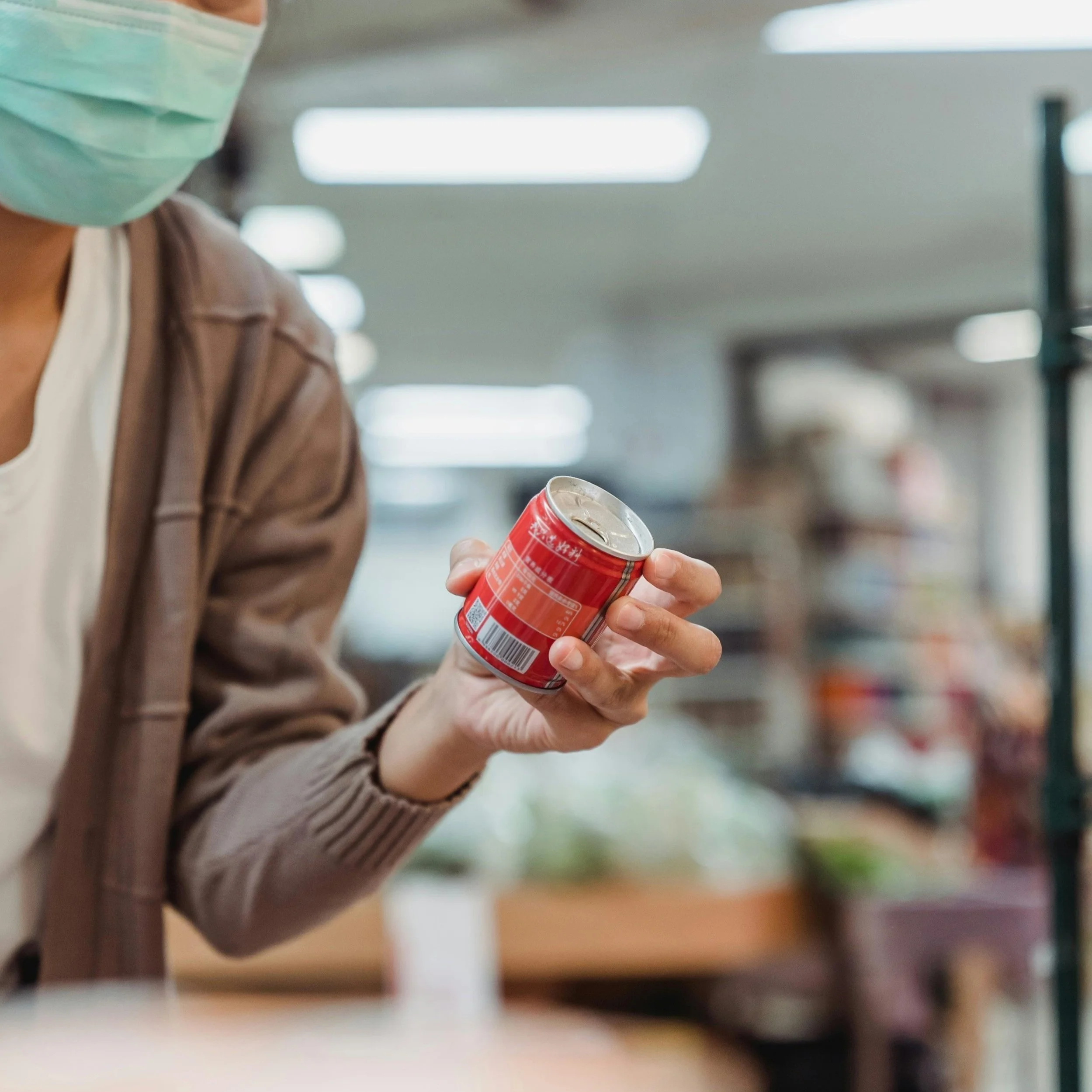 Person wearing a face mask holding a small red canned beverage in a store aisle.