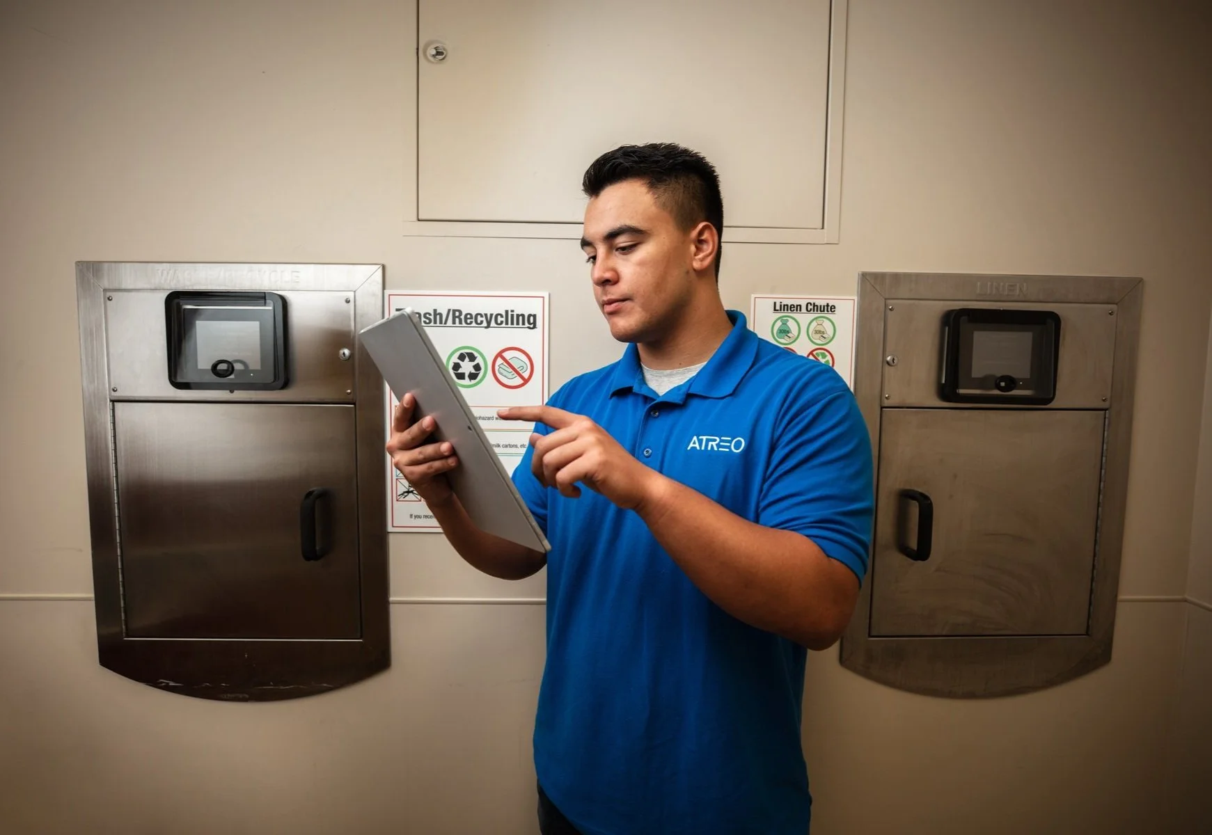 A man in a blue polo shirt standing in front of recycling and linen chute receptacles, looking at a tablet device.