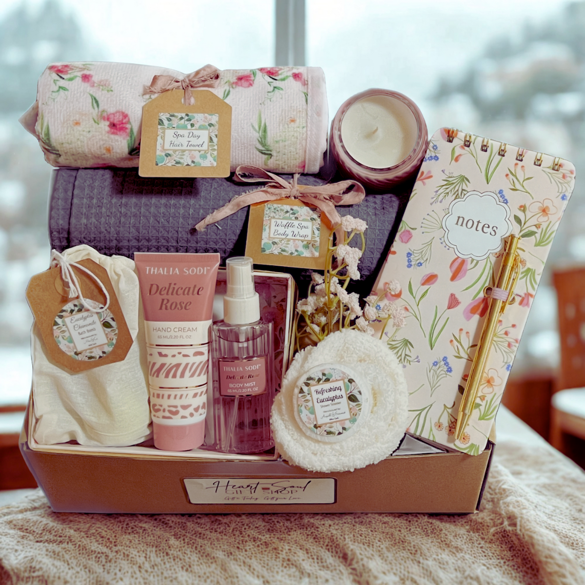 Gift box containing spa and relaxation items, including a floral towel, body wrap, scented candle, notepad with floral cover, hand cream, body mist, bath bomb, and a gold pen.