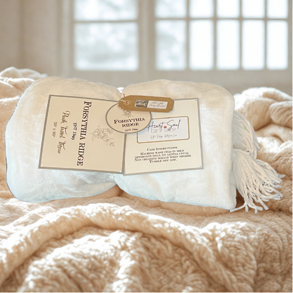 A cream-colored pillow with a tag and paper label, resting on a textured beige blanket in front of a window with curtains.