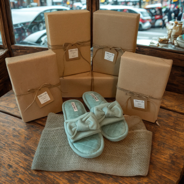 Five neatly wrapped gift boxes with brown paper and tied with string, a pair of light blue slippers with bows, and a piece of burlap fabric on a wooden surface, with a window and parked cars visible outside.