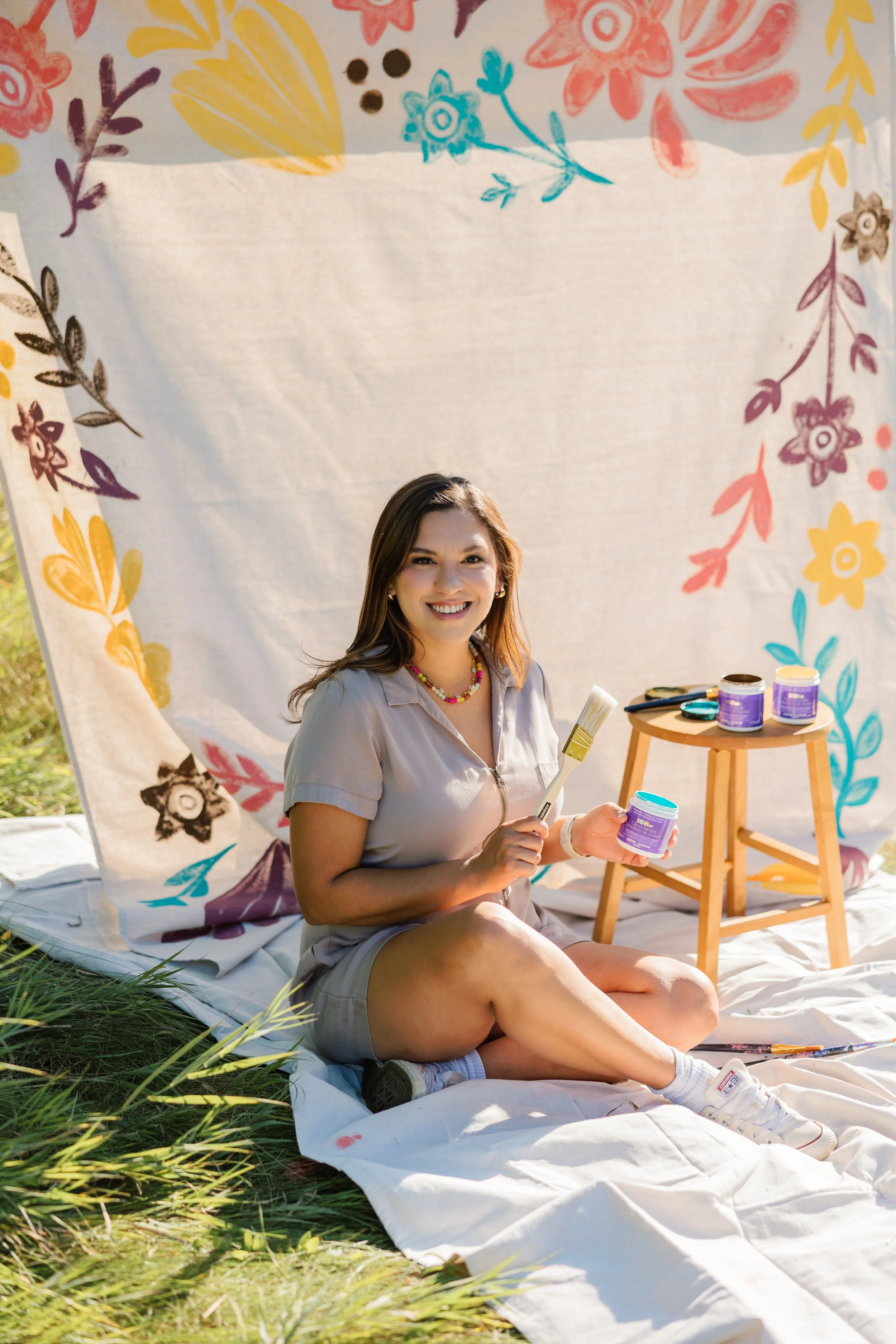 A young woman sitting outdoors on a white blanket, holding a jar of paint and a paintbrush, surrounded by jars of paint, with a decorative floral fabric backdrop.