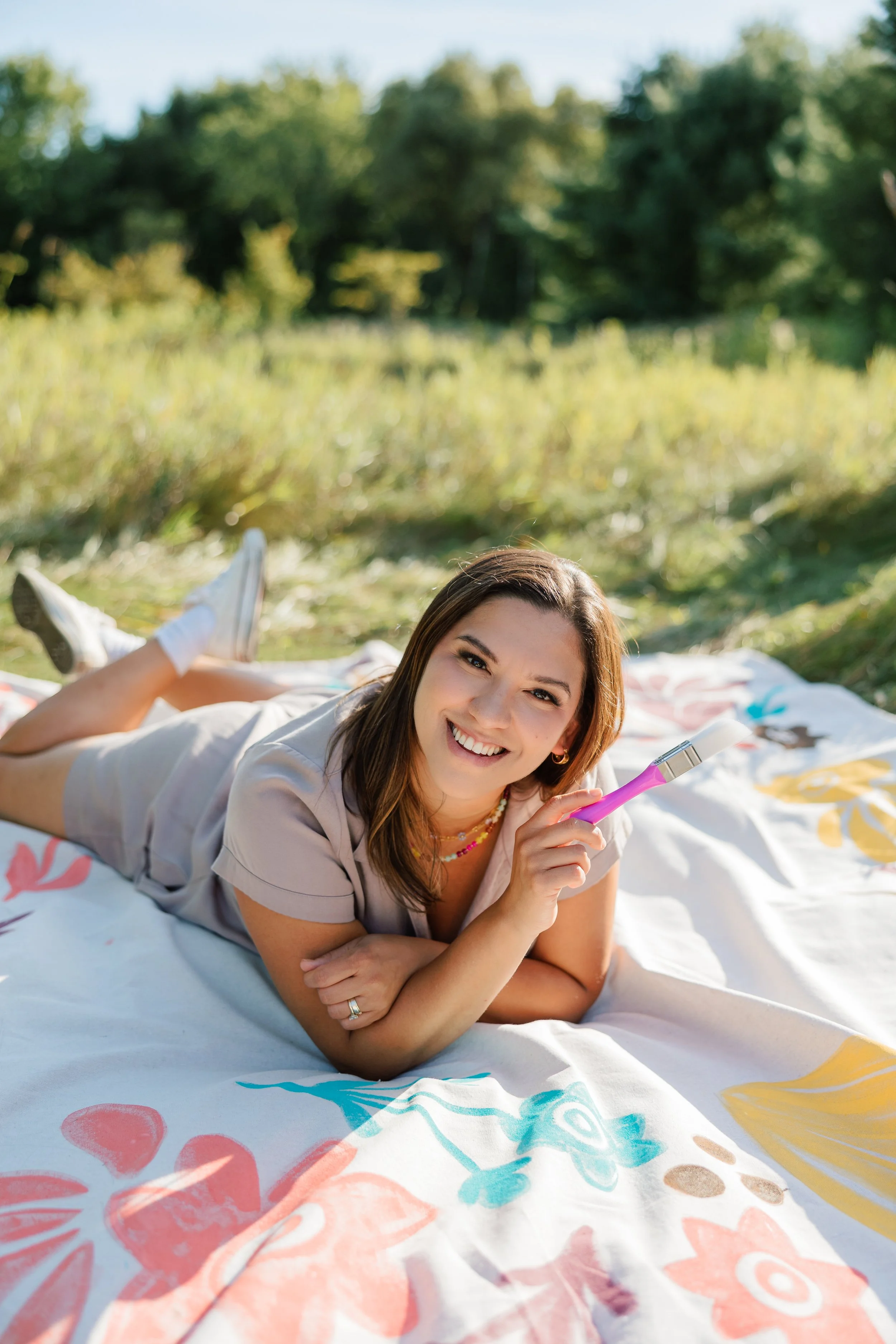 A woman lying on her stomach on a colorful floral blanket outdoors, smiling and holding a paintbrush. She is in a grassy area with trees in the background on a sunny day.