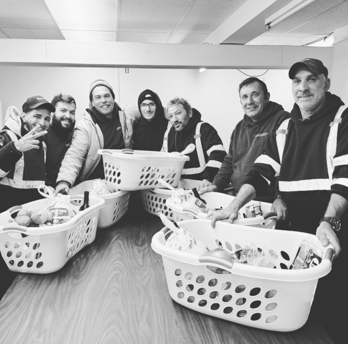 Group of eight men standing behind a table with laundry baskets filled with knitted stuffed animals, smiling and posing for the camera in an indoor setting.
